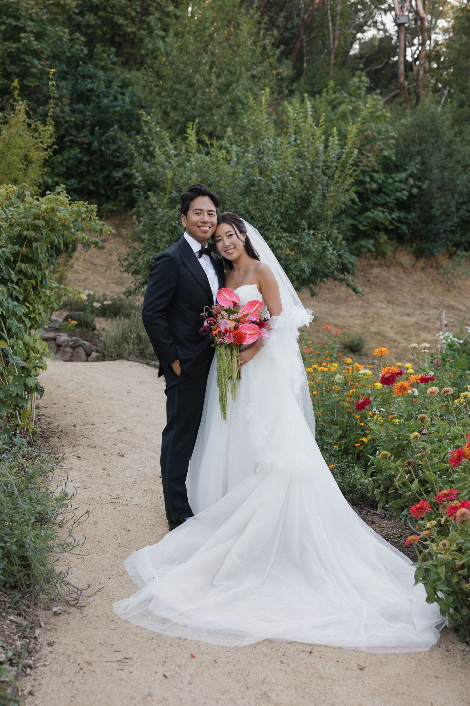 Newlyweds pose together on a garden path surrounded by vibrant summer flowers.