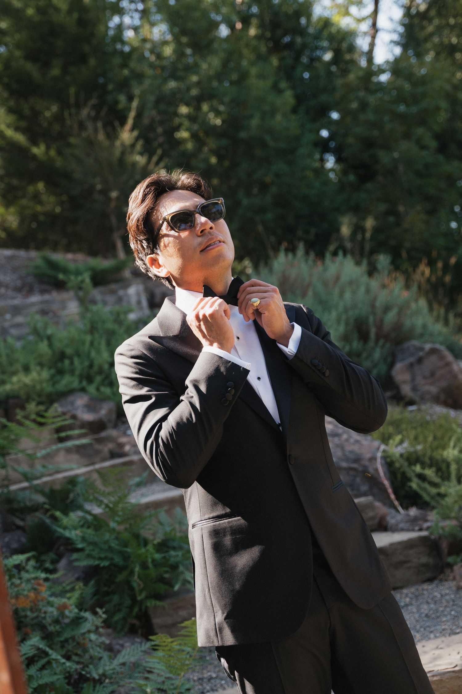 A person in a black suit and sunglasses poses outdoors in a garden setting.