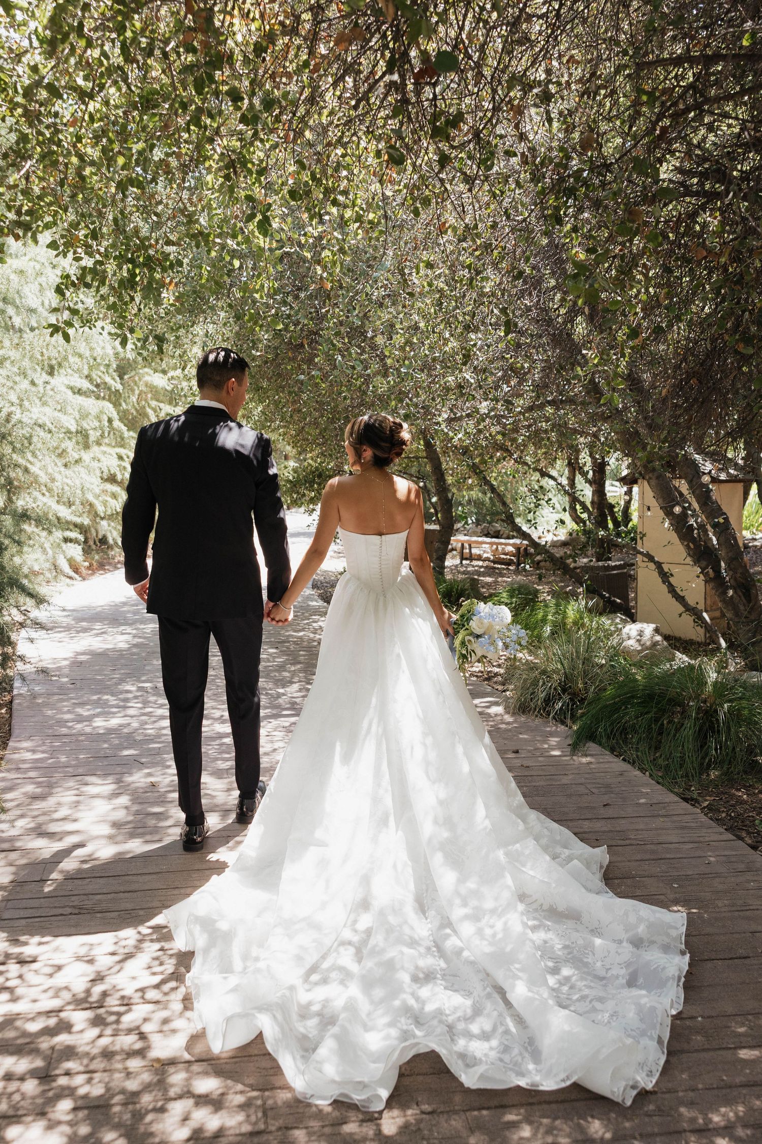 A bride in a flowing white gown and groom walking along a tree-lined garden path after their wedding ceremony.