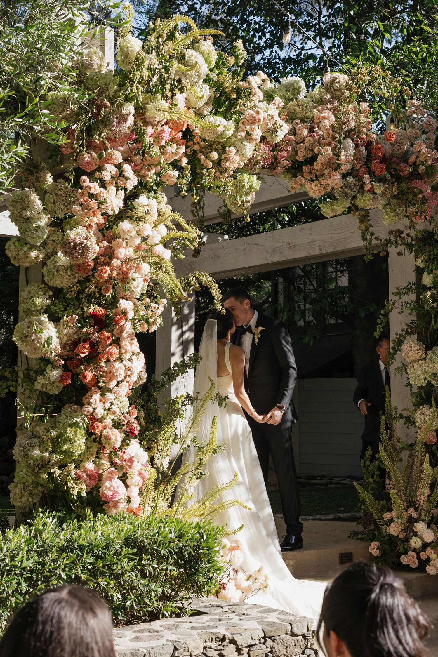 Bride and groom share a tender moment under a lush floral arch adorned with pink and white blooms at an outdoor wedding ceremony.