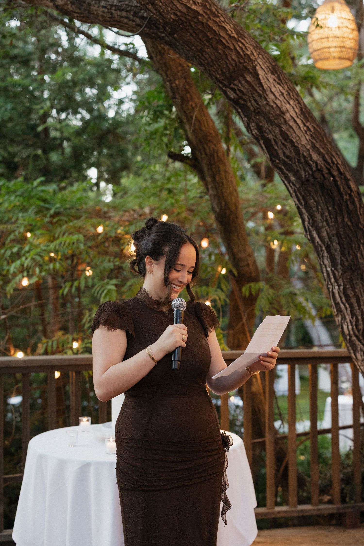 Smiling pregnant woman in brown dress giving a speech at an outdoor wedding reception surrounded by string lights.