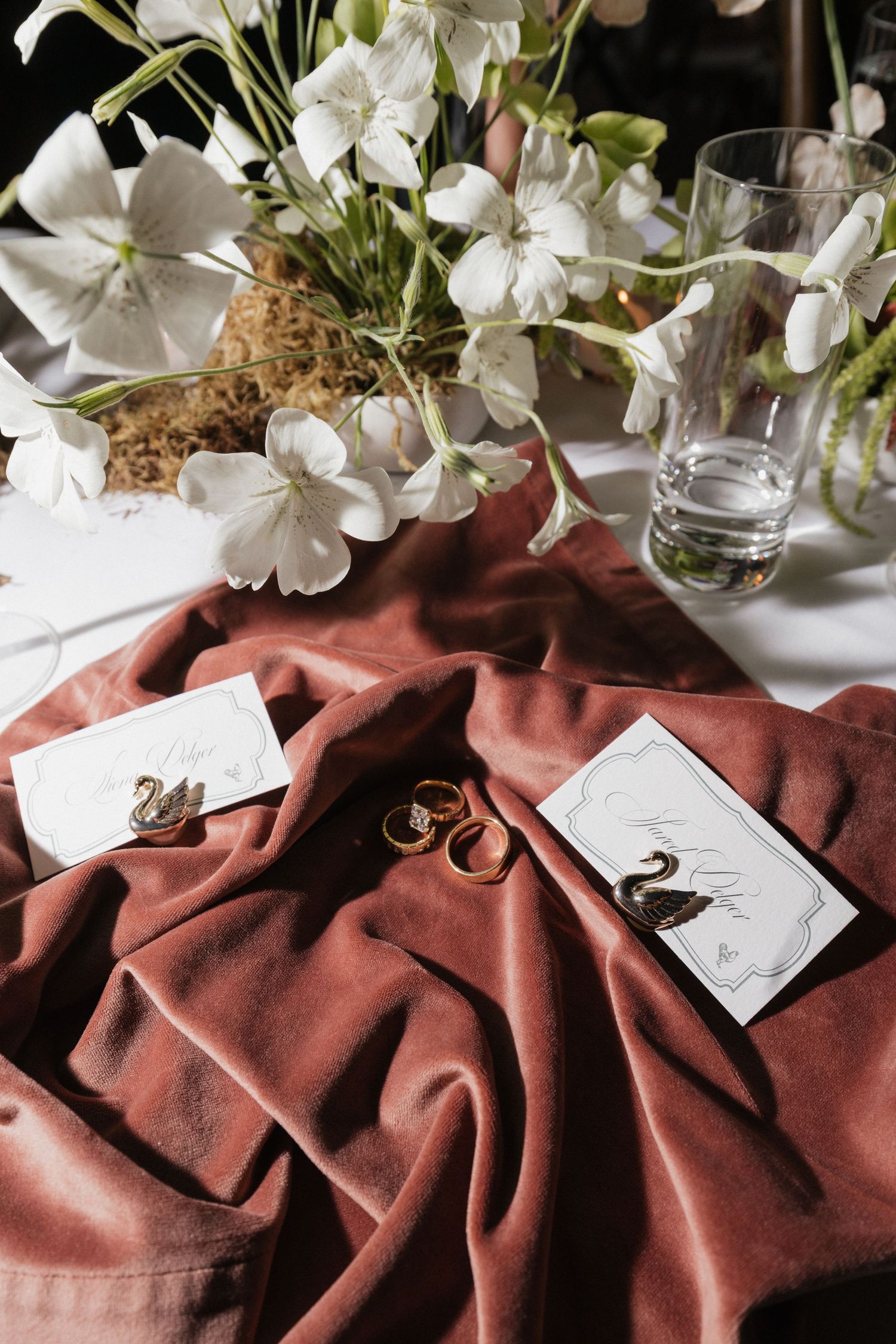 Elegant wedding flat lay with gold rings, swan place cards, and white florals on dusty rose velvet fabric.