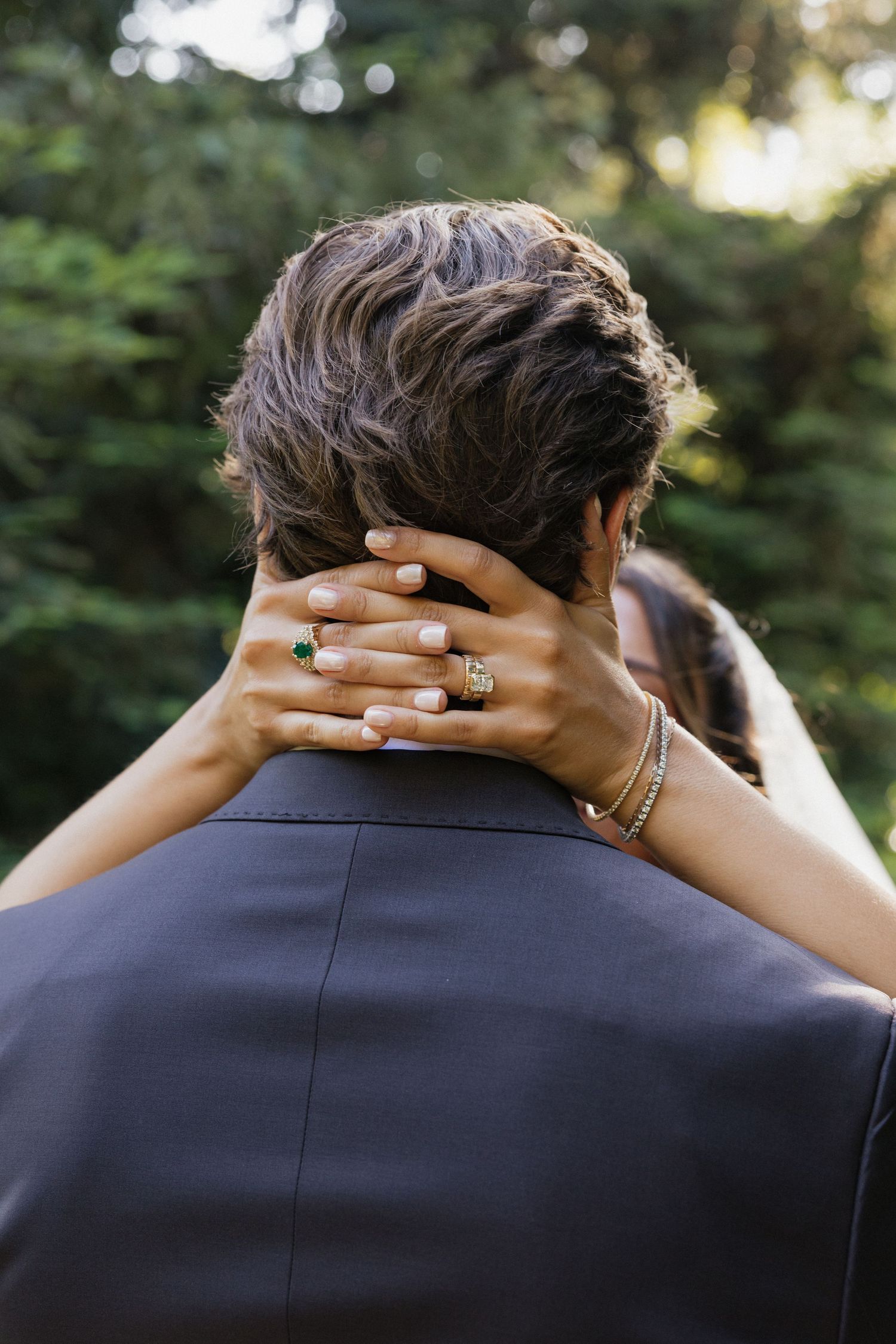 Bride placing hand on grooms neck showing emerald gold ring and bracelet at outdoor wedding.