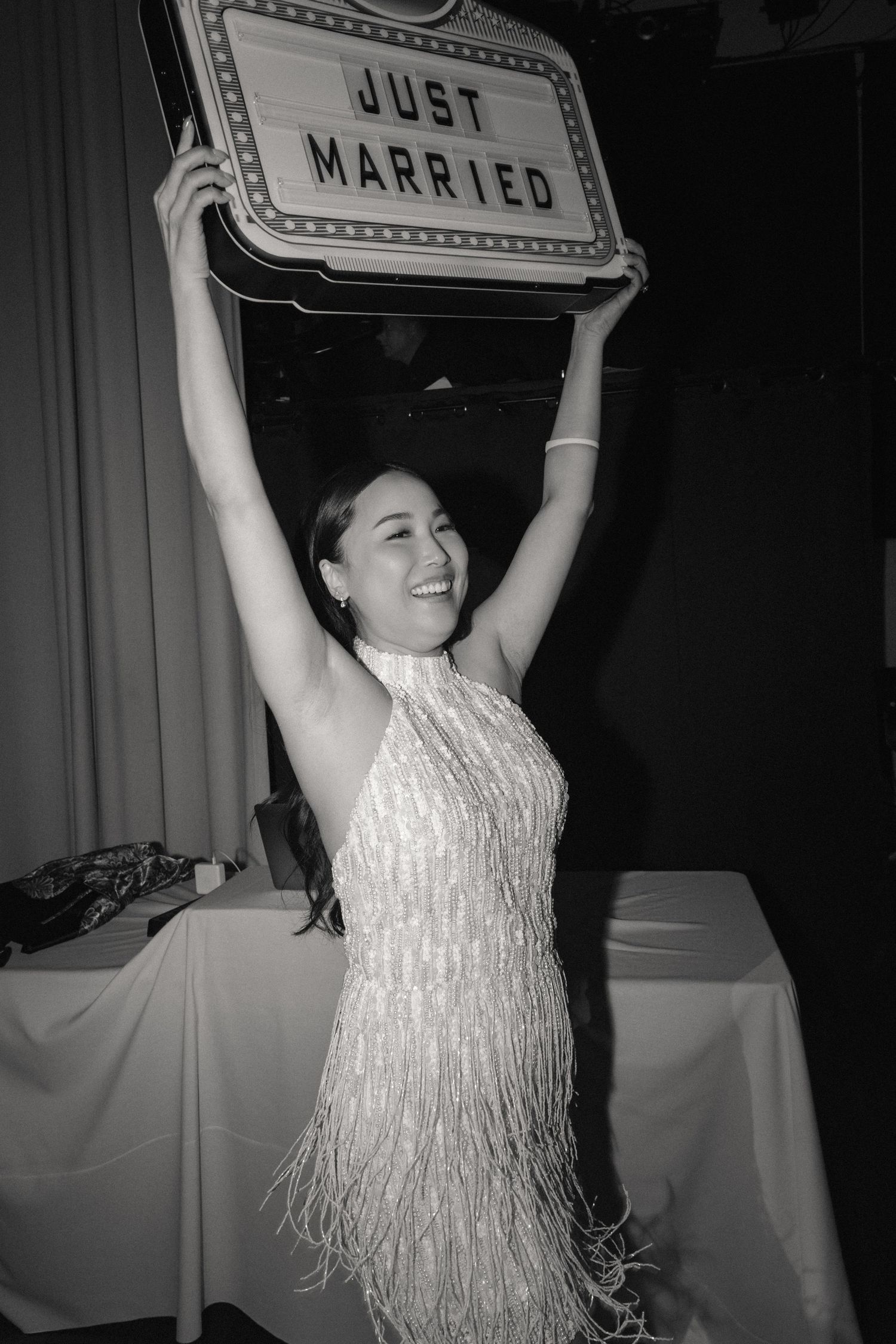 Black and white photo of a smiling bride in a fringed dress holding a Just Married sign above her head.