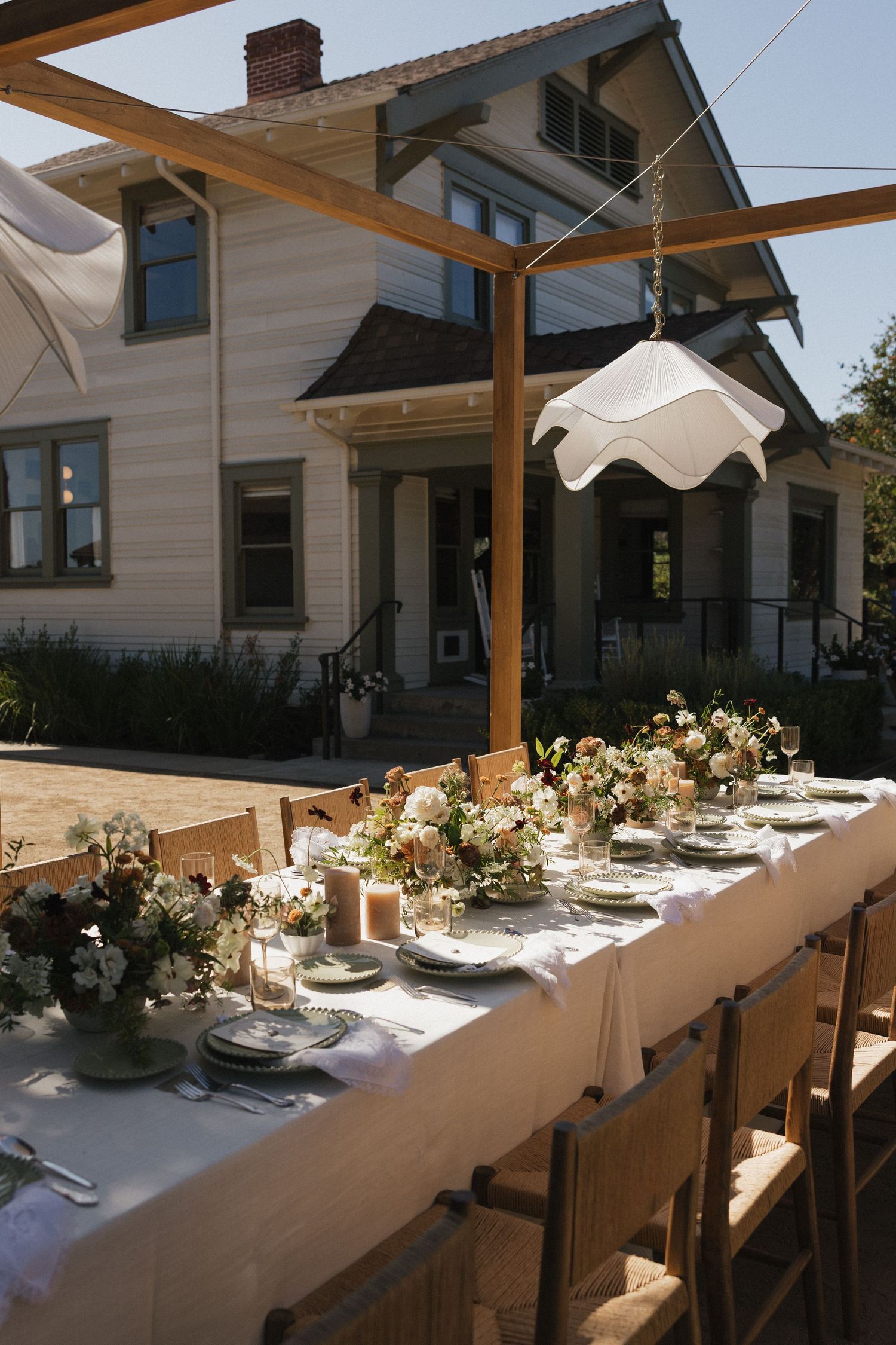 Elegant outdoor dining table setup with white linens and floral centerpieces under a covered patio area.