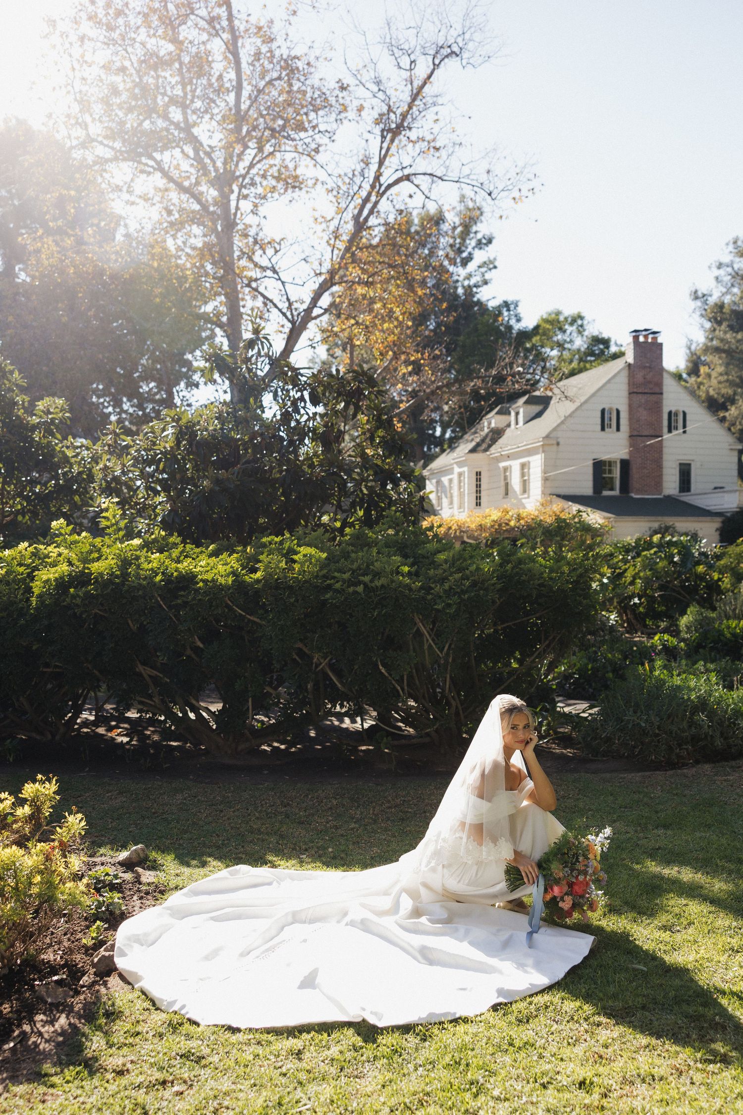 A wedding dress with a long flowing train spreads across a sunlit lawn with white houses in the background.