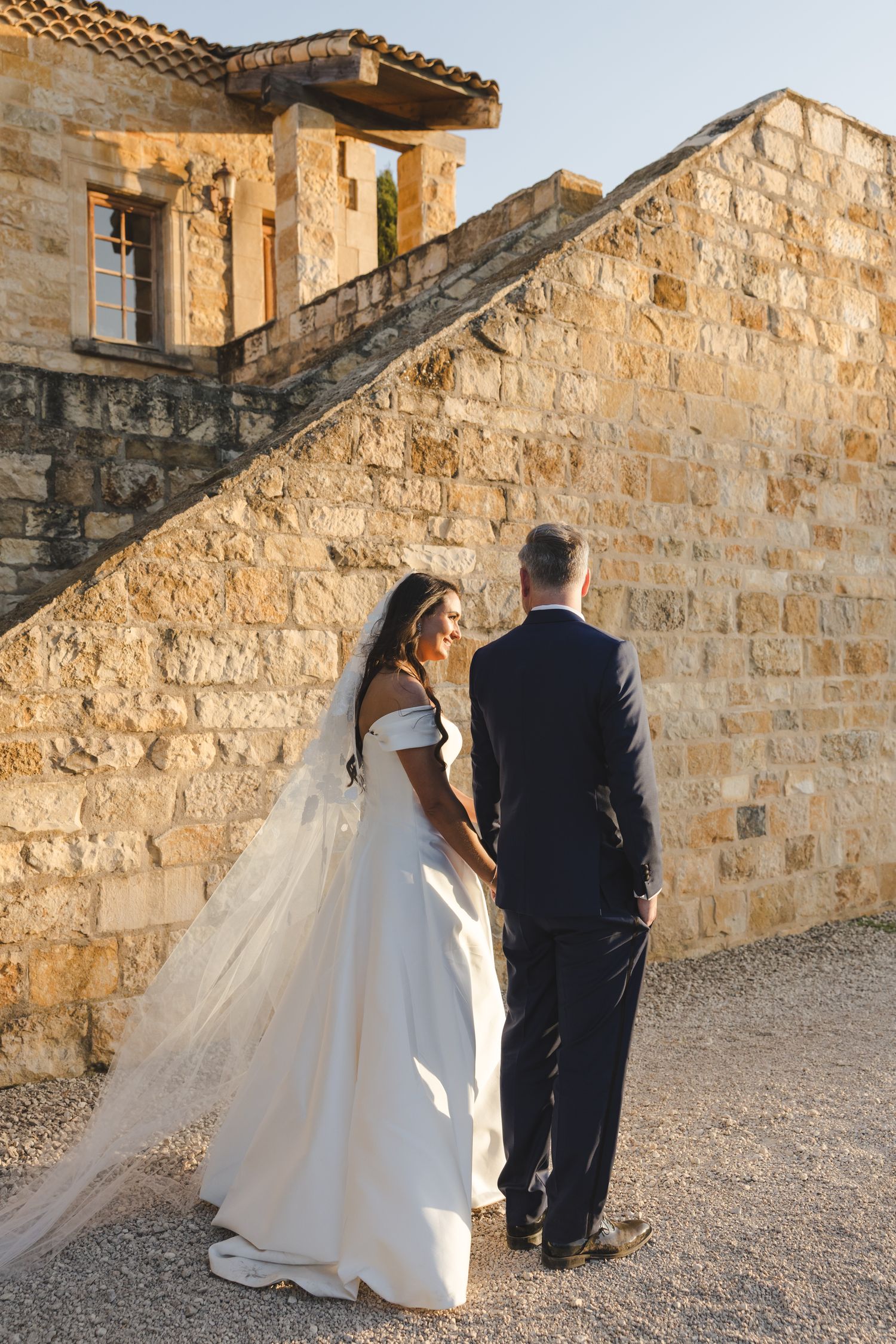 Wedding couple standing against rustic stone wall architecture at sunset.