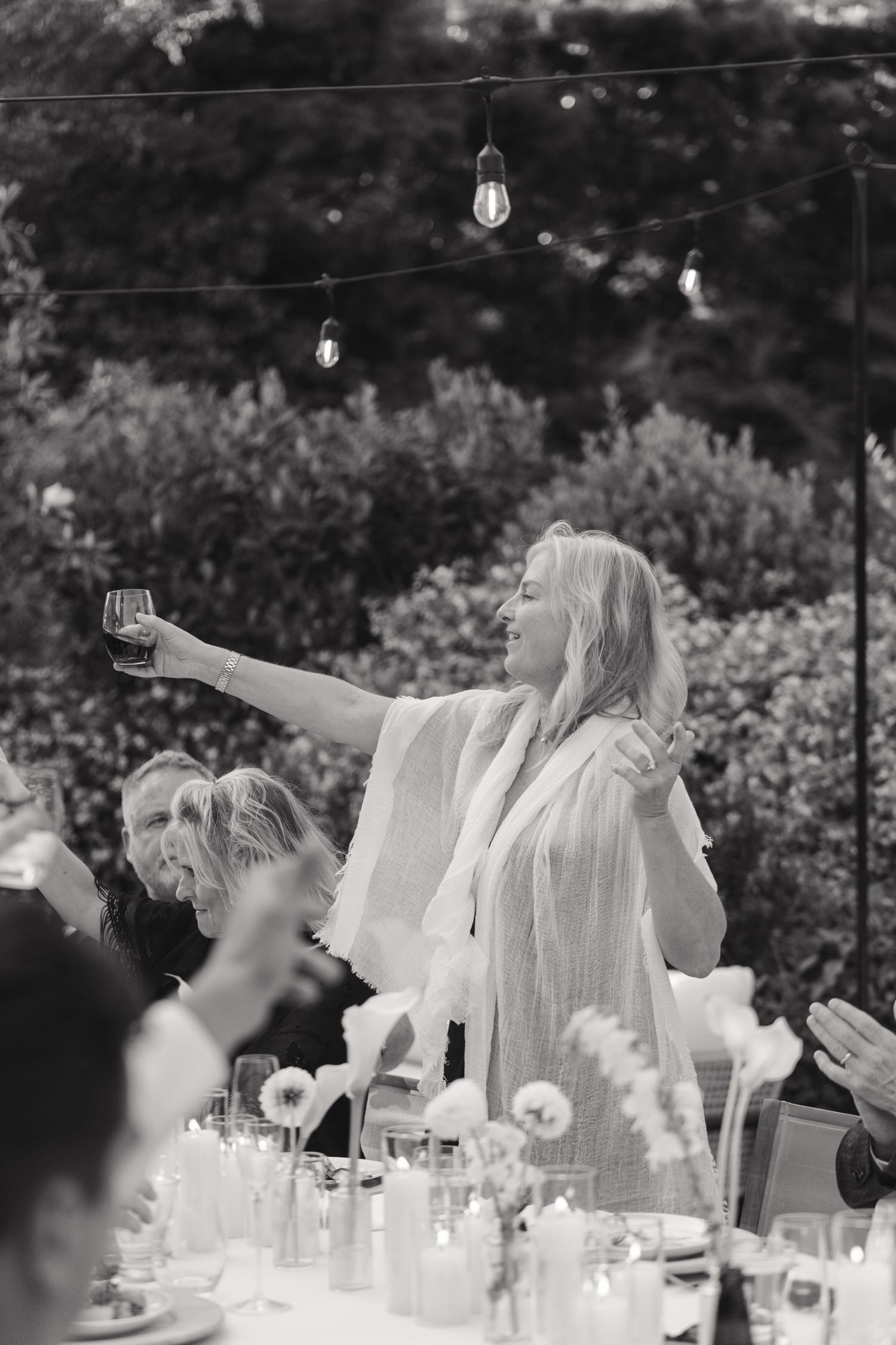 A celebratory outdoor dinner scene with raised wine glass under string lights, captured in black and white.