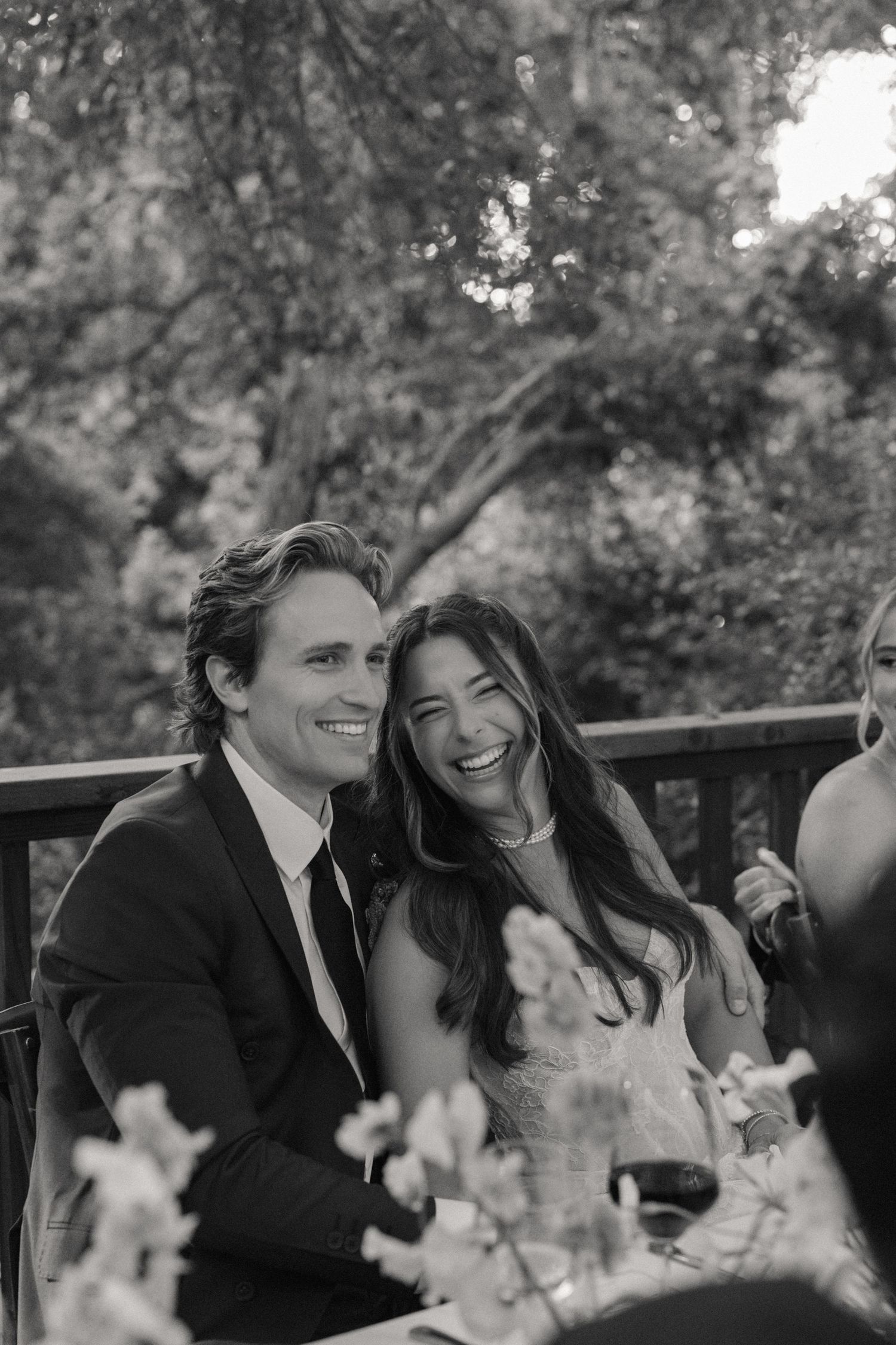 Black and white photo of a laughing couple at an outdoor wedding reception seated at a floral table.