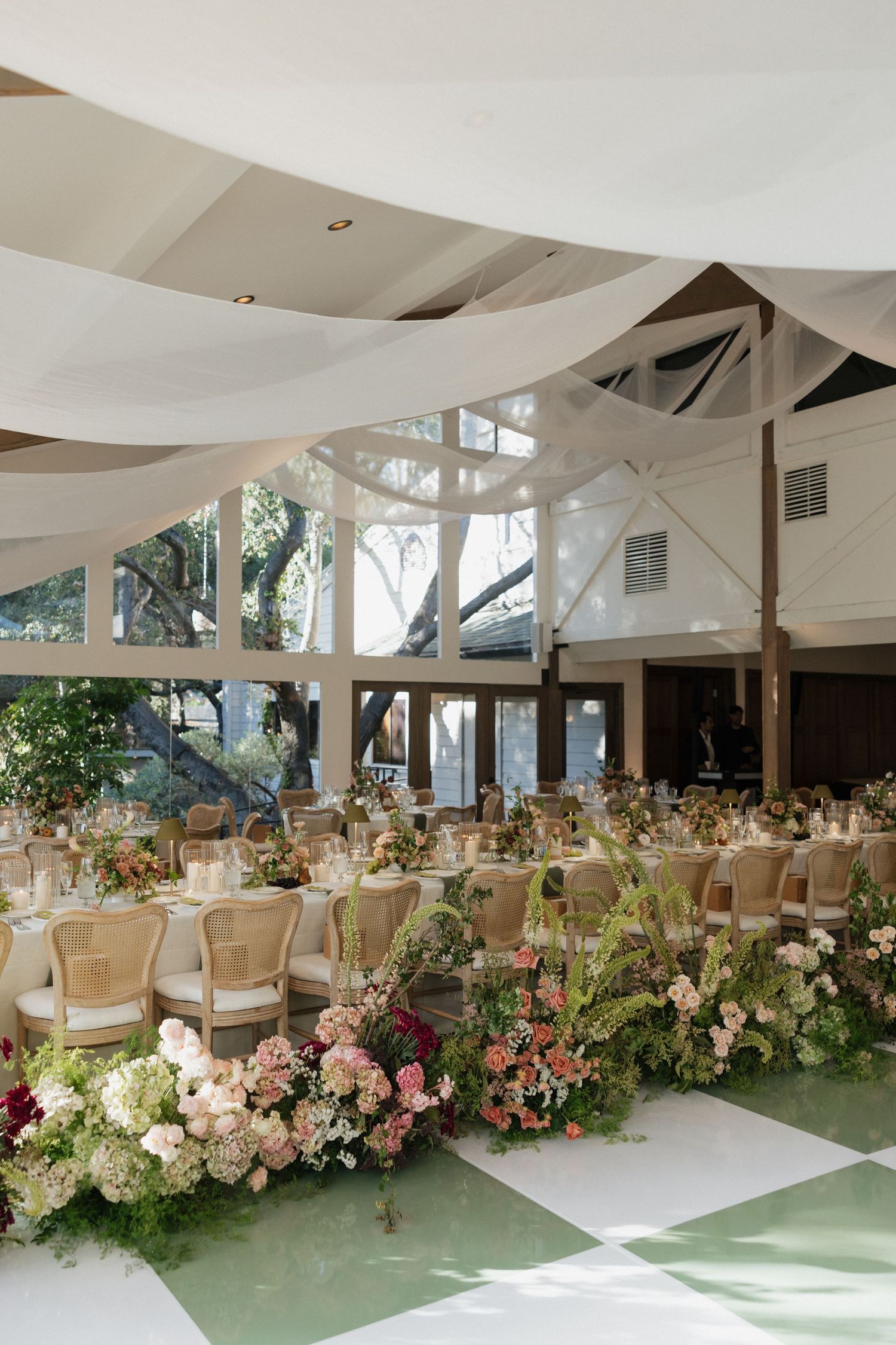 Elegant wedding reception with draped white fabric ceiling, floral arrangements, and checkered floor in a bright venue.