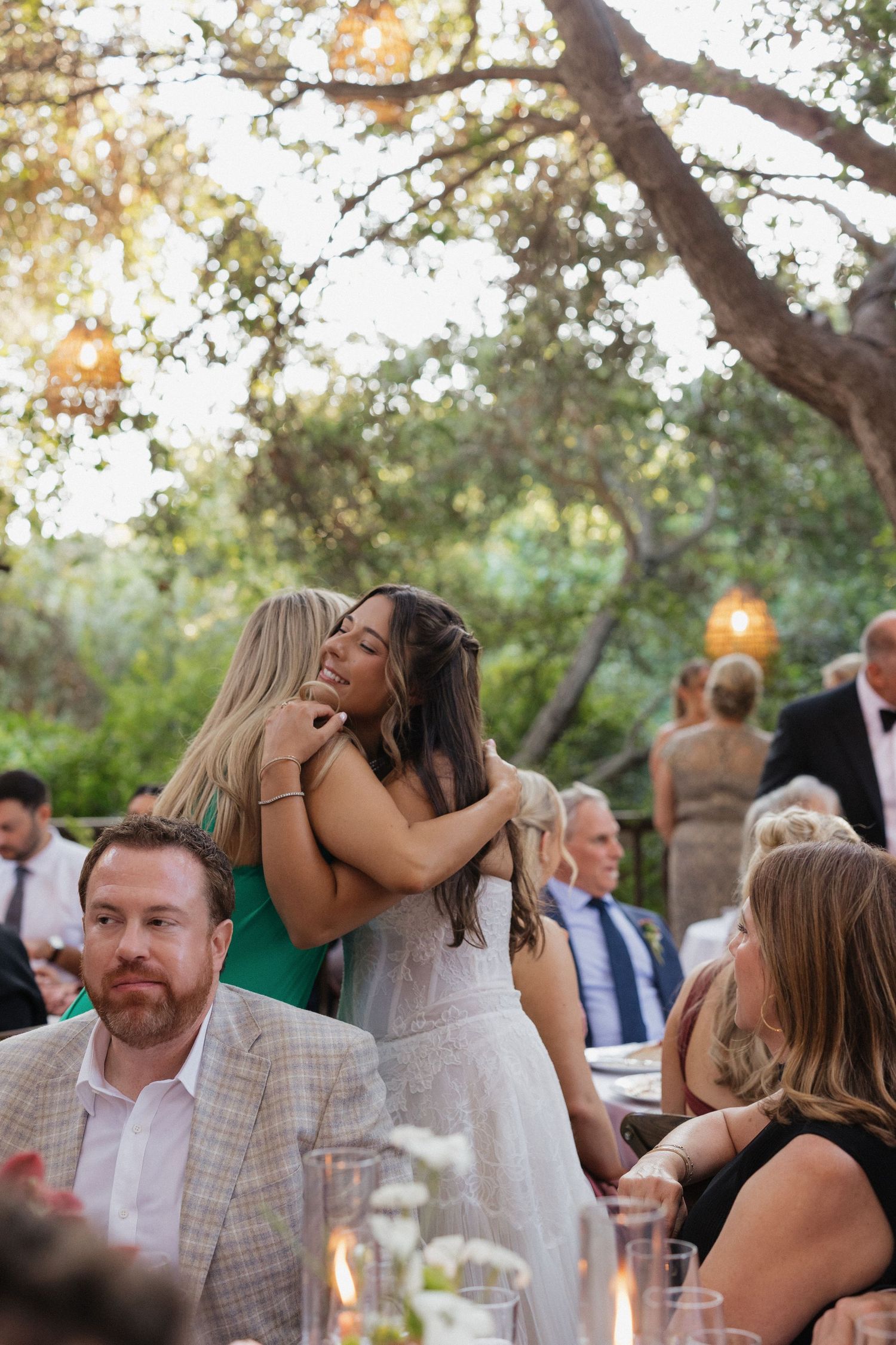 Bride shares warm embrace with guest at outdoor wedding reception under canopy of trees.