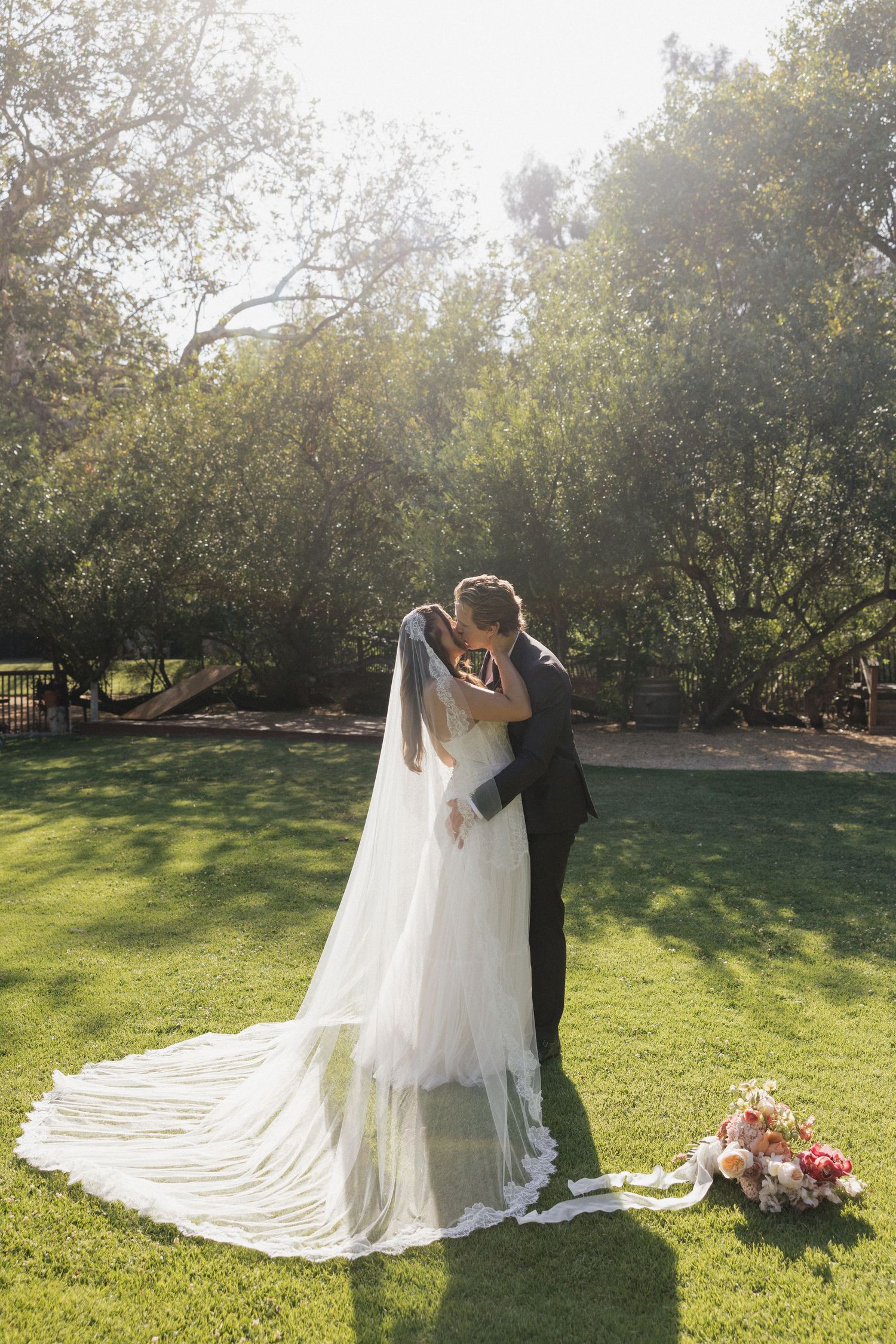 Bride and groom share romantic kiss on sunlit green lawn, brides cathedral veil trailing behind.