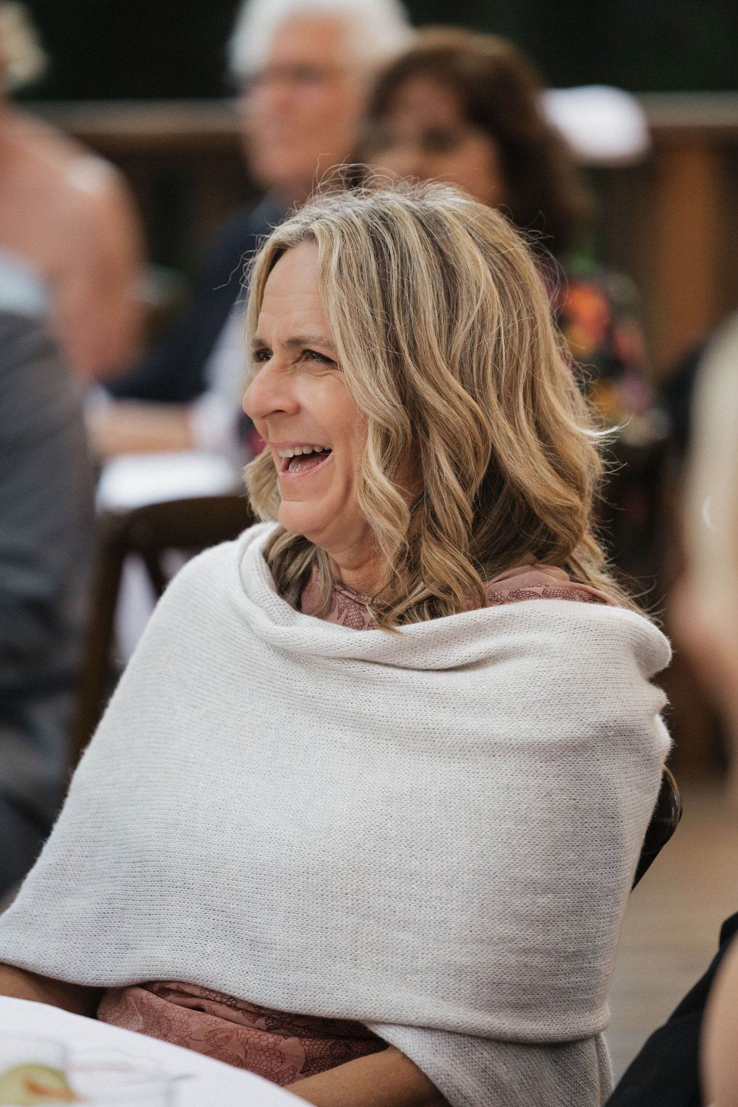 Joyful female wedding guest laughs while seated outdoors, wearing a cozy gray wrap shawl.