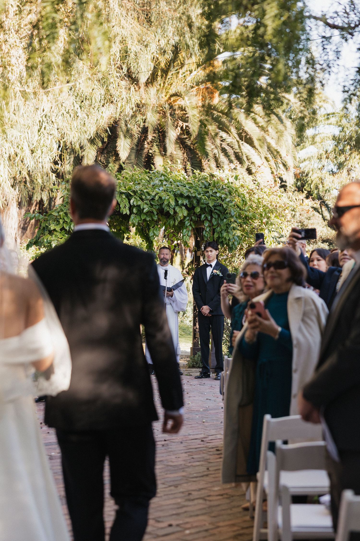 Wedding ceremony taking place on a brick pathway under hanging moss trees with guests in attendance.