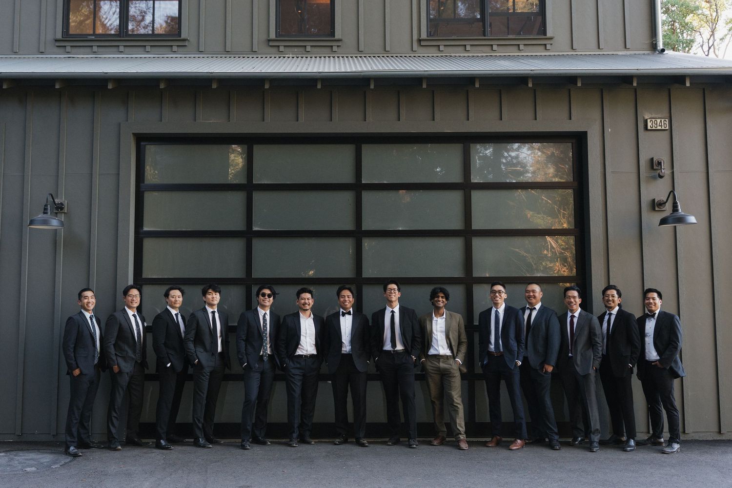 A line of groomsmen in black suits pose against a grey building exterior.