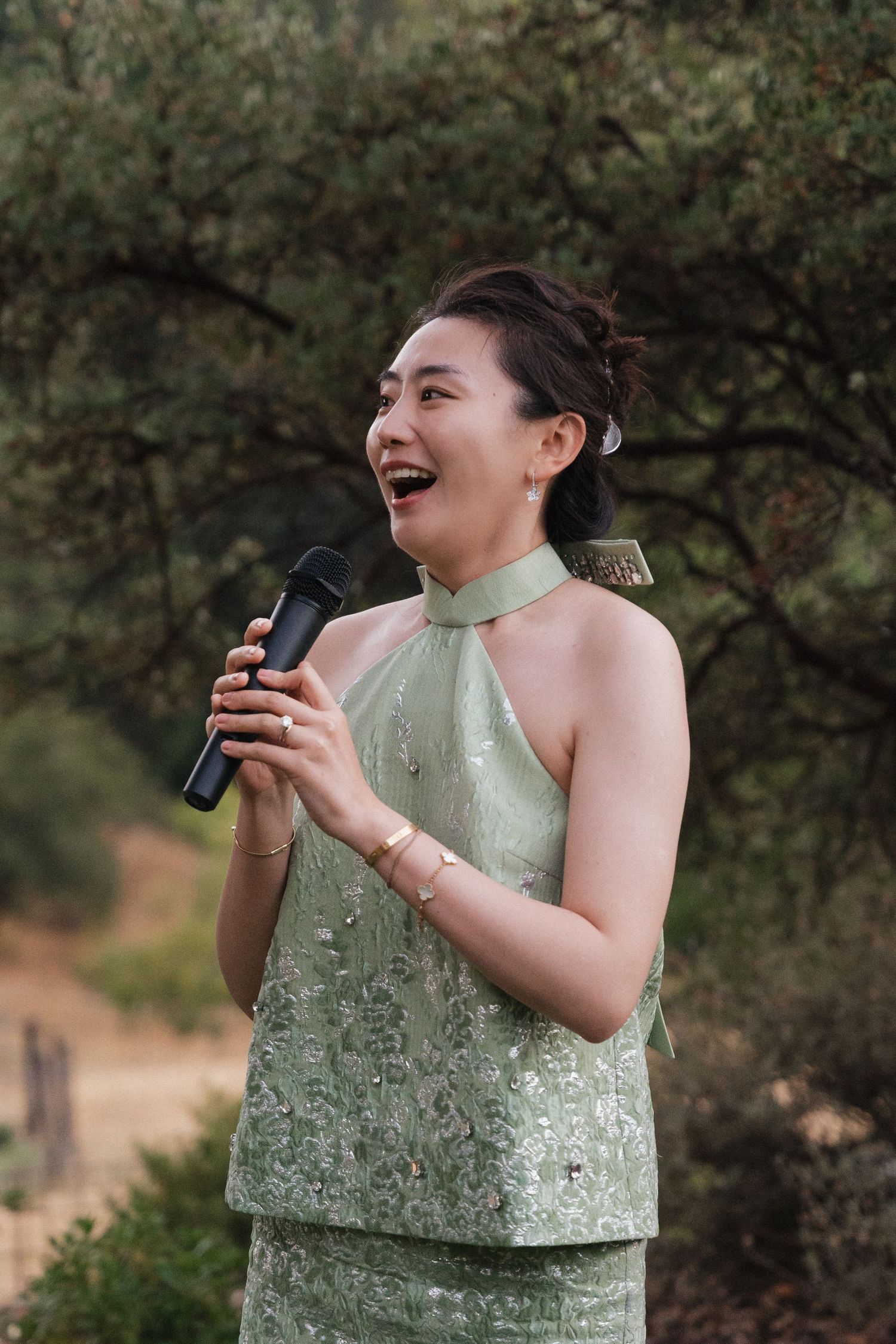 A woman in a mint green halter dress speaks into a microphone while laughing outdoors.
