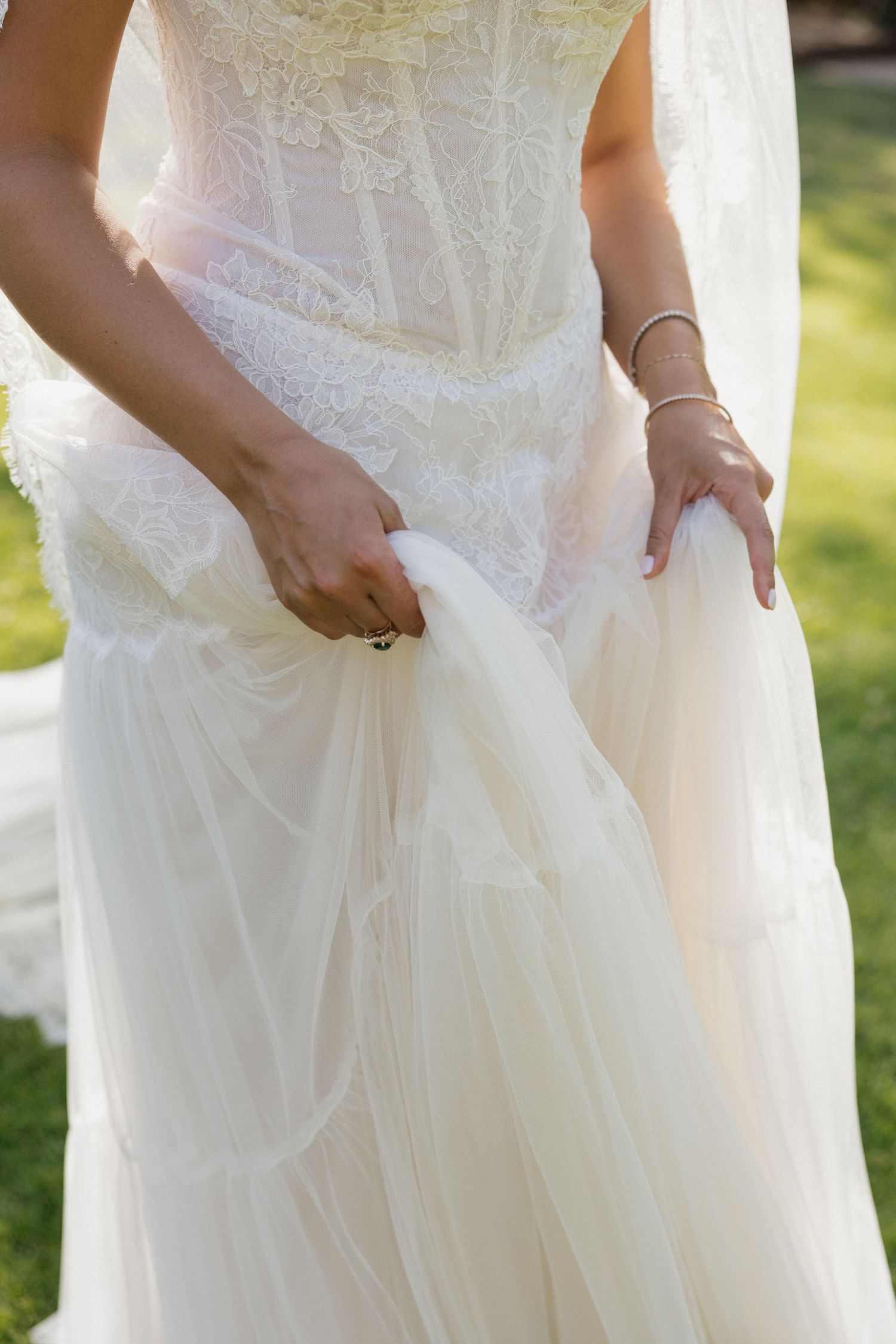 Bride in elegant white lace and tulle wedding dress holding skirt outdoors on green lawn.