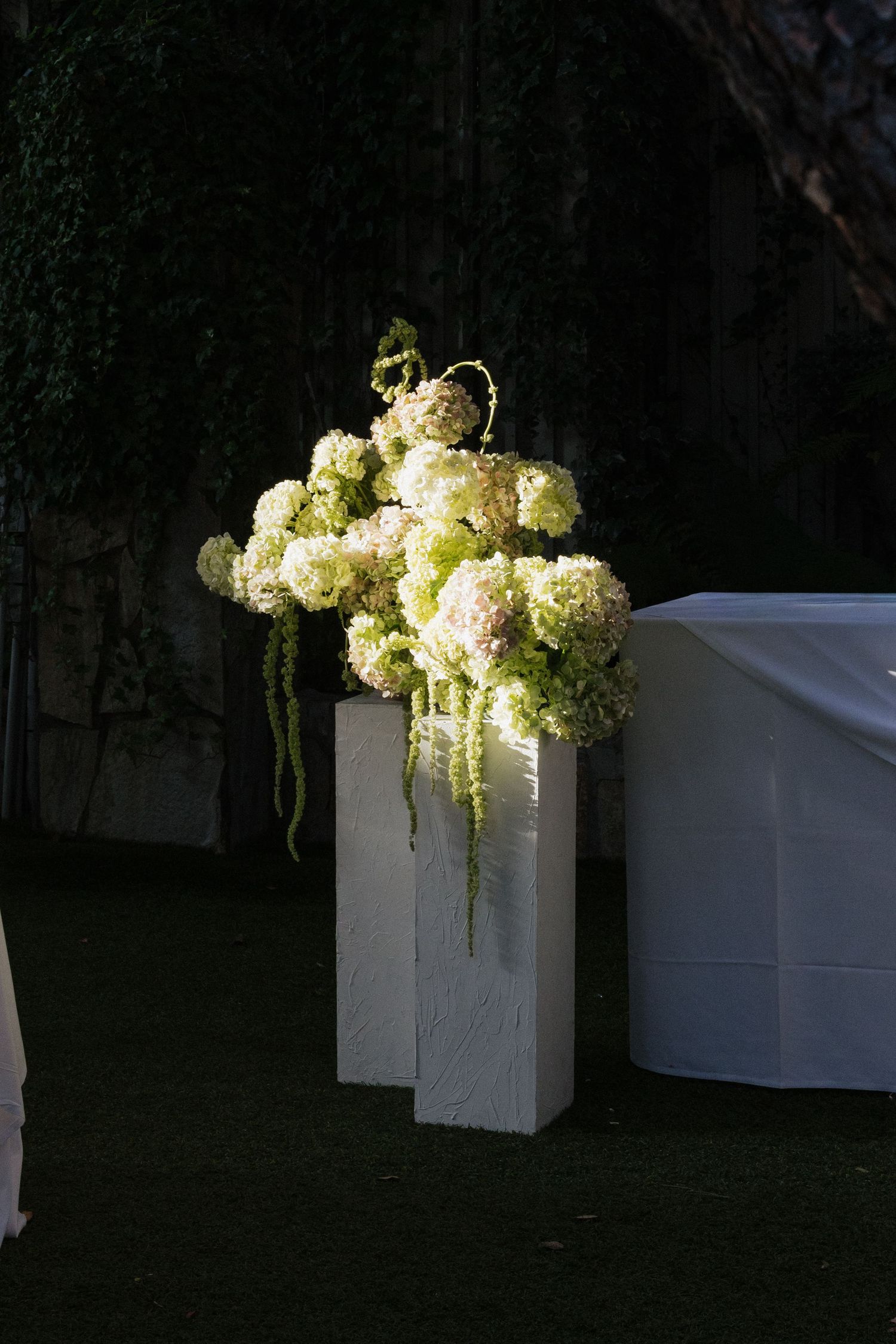 White hydrangea and green floral arrangement on tall white pedestal in dark garden wedding ceremony setting.