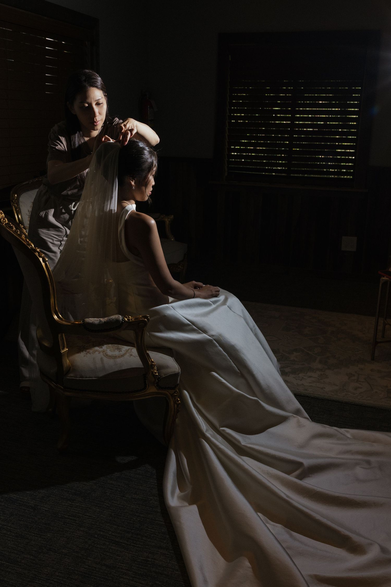 Bride in elegant white gown seated in ornate chair while bridesmaid attaches veil in dimly lit room.