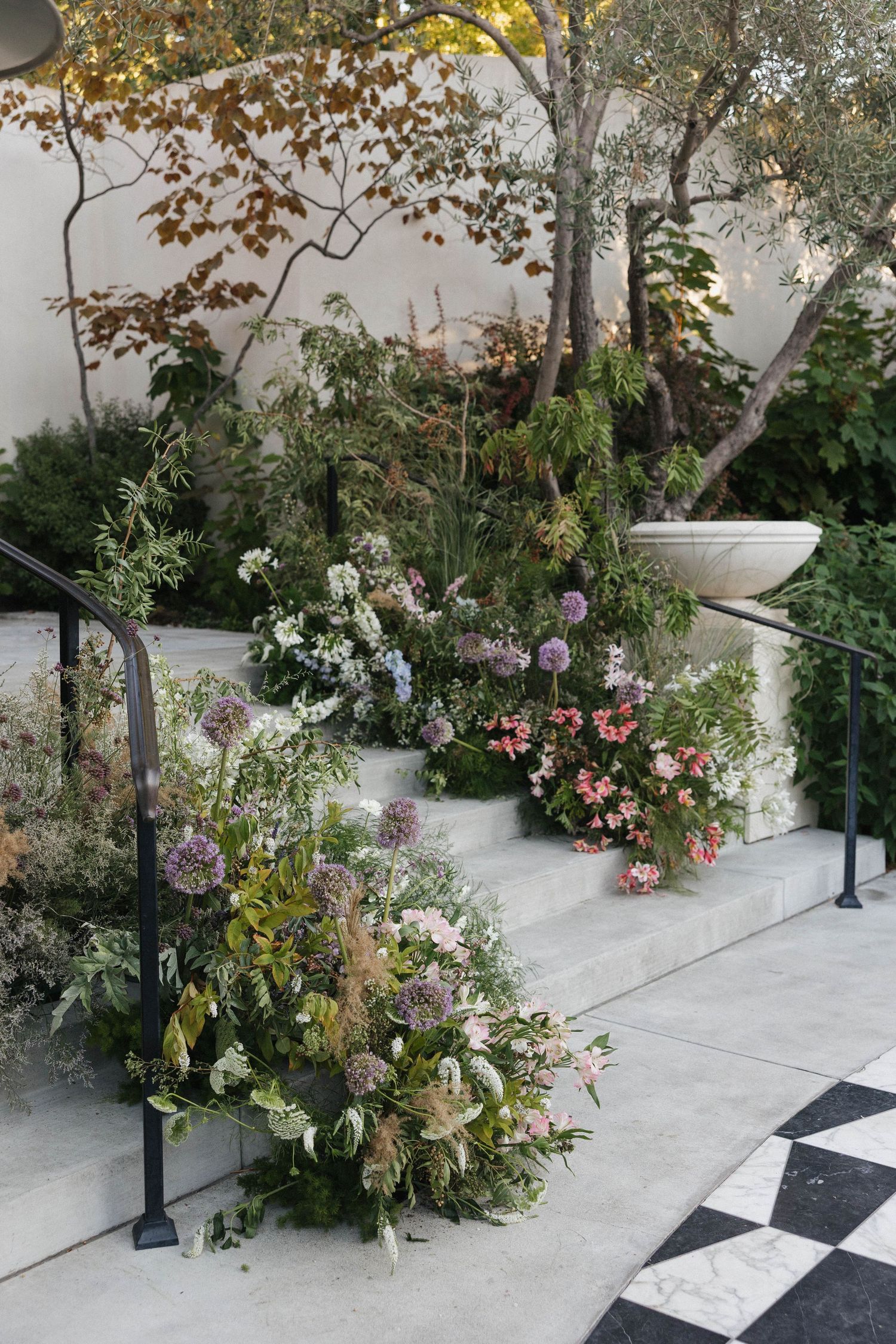 Outdoor garden path with pink flowers and greenery alongside white stone walkway featuring decorative lampposts.