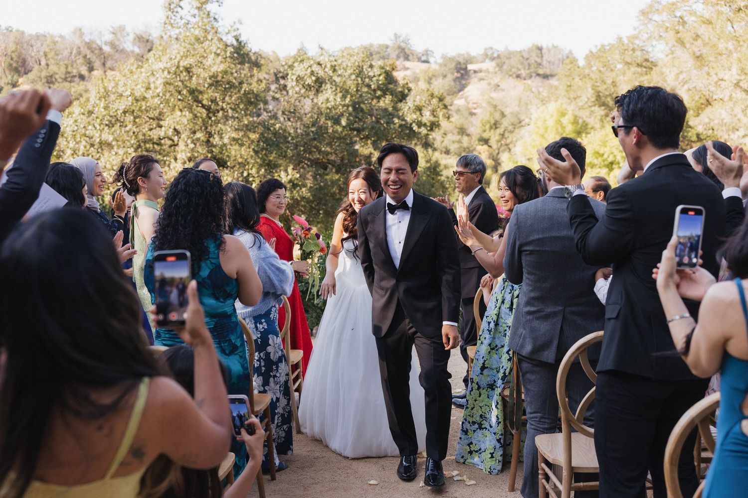 Wedding party and guests mingle outdoors under a canopy of trees during a daytime celebration.