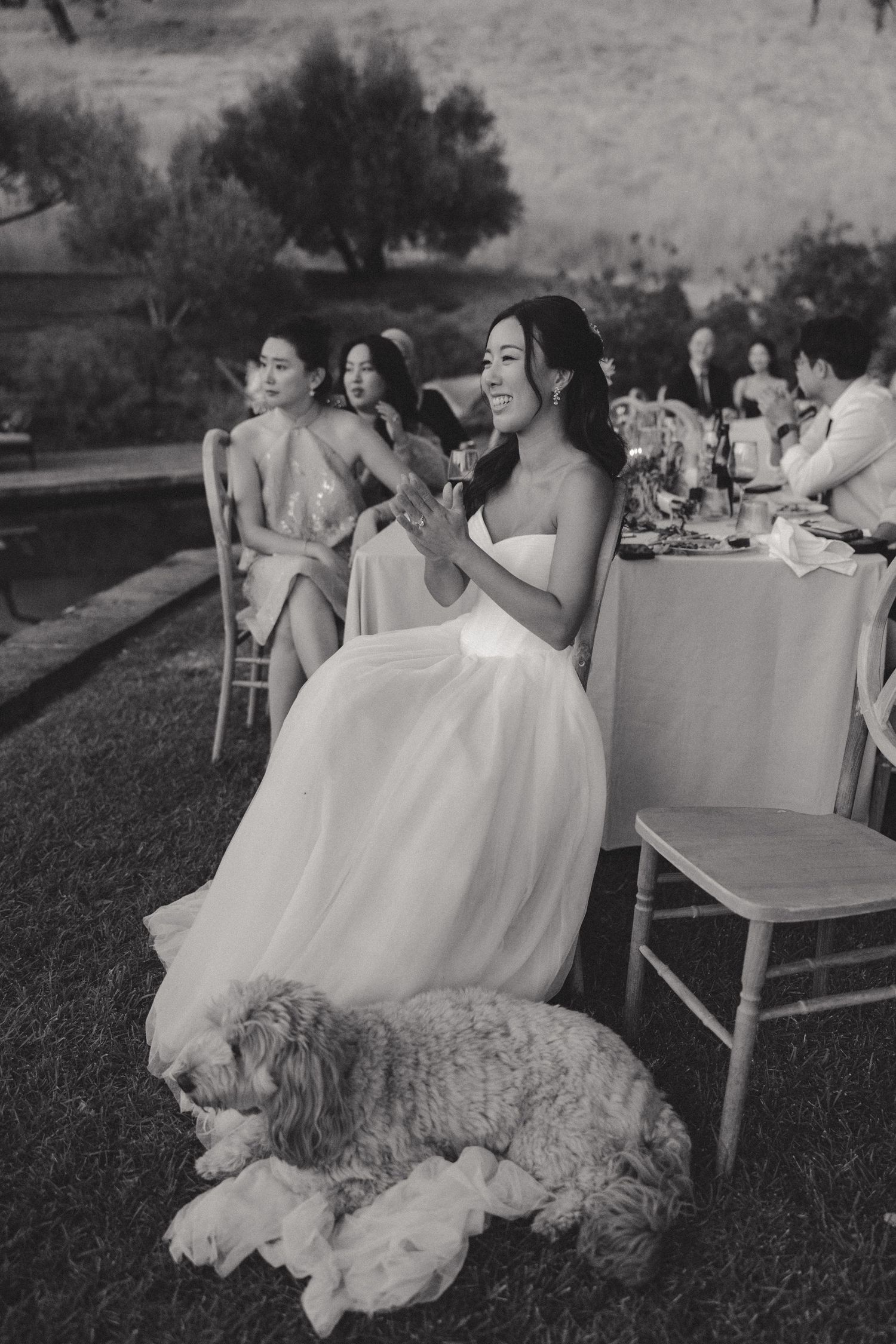 Wedding party members laughing while seated at outdoor reception tables in black and white.