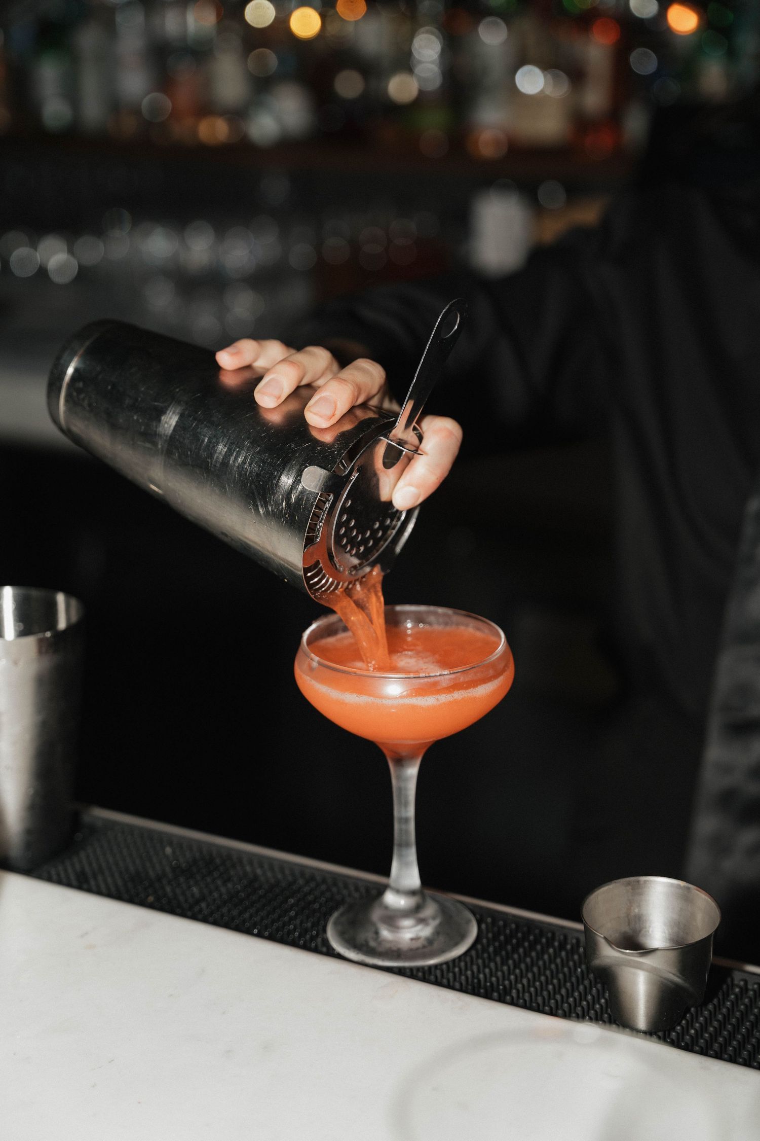 Bartender pouring orange cocktail into coupe glass at dimly lit bar.