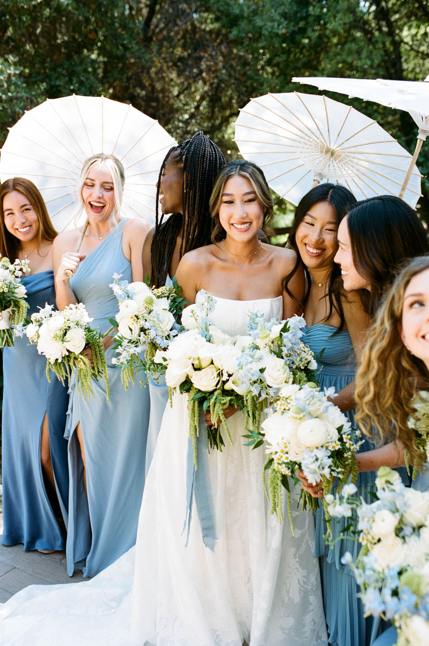 Wedding party in light blue bridesmaid dresses holding white parasols and floral bouquets.