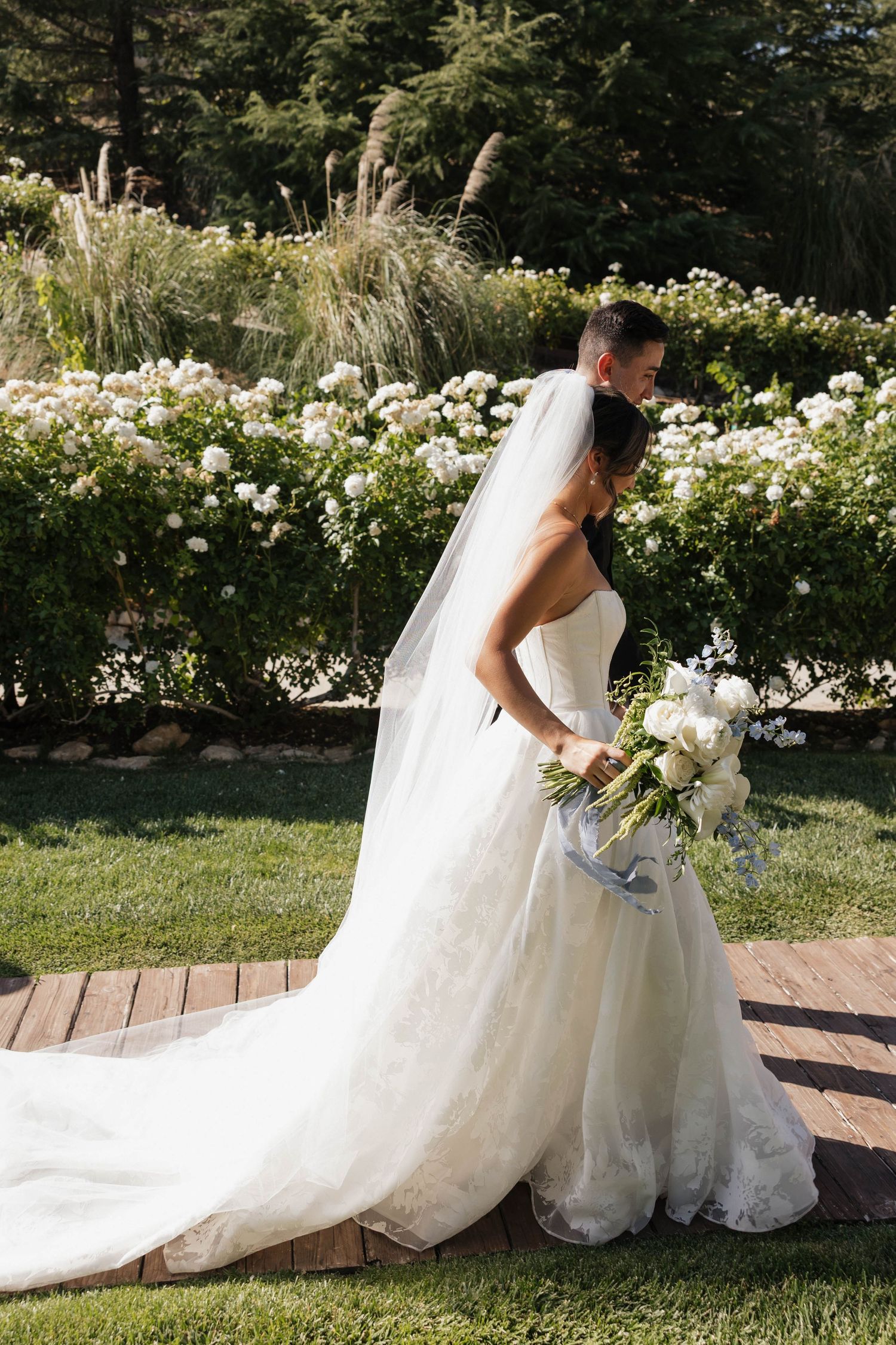 Wedding dress train elegantly arranged on grass near blooming white garden flowers.