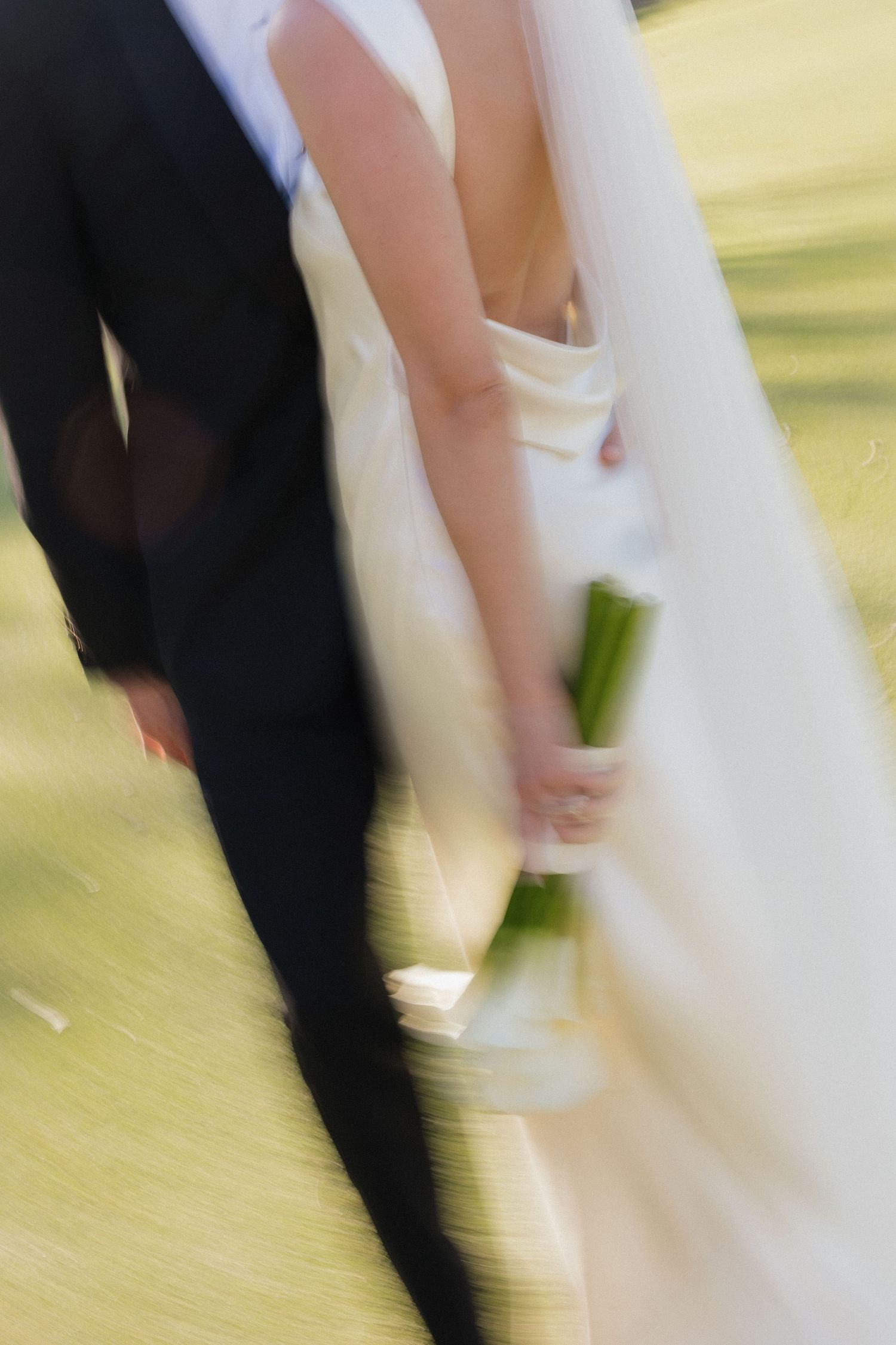 Blurred motion photo of bride in white gown and groom in black suit holding hands and walking together outdoors.