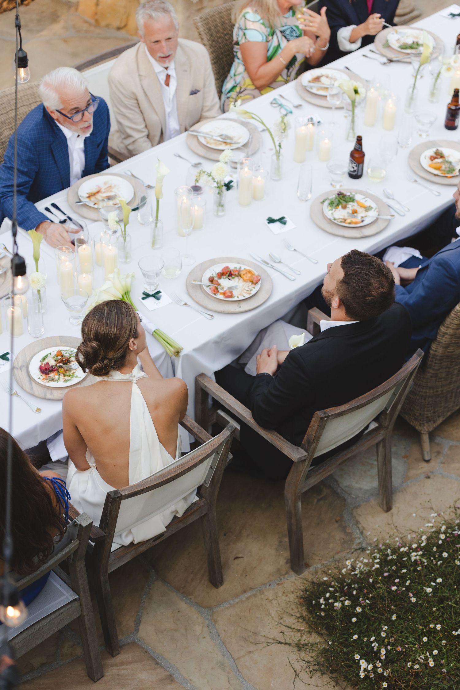 Overhead view of an elegant outdoor wedding reception dinner with guests seated at a long white-clothed table.