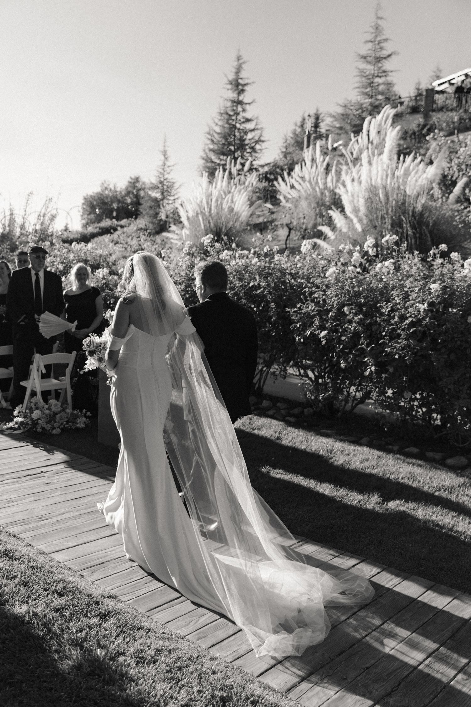 Wedding party walking down a garden path in elegant white and dark attire with flowing veils.