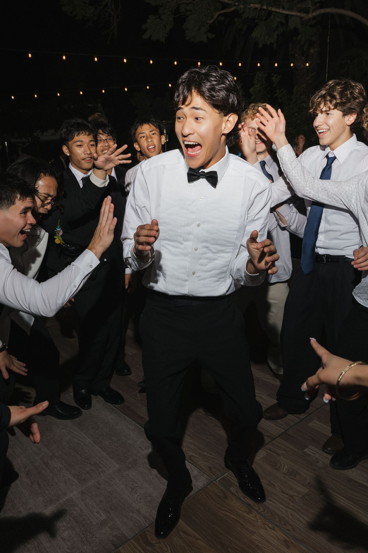 Wedding guests in formal attire dance energetically in a line on a dark wooden dance floor.