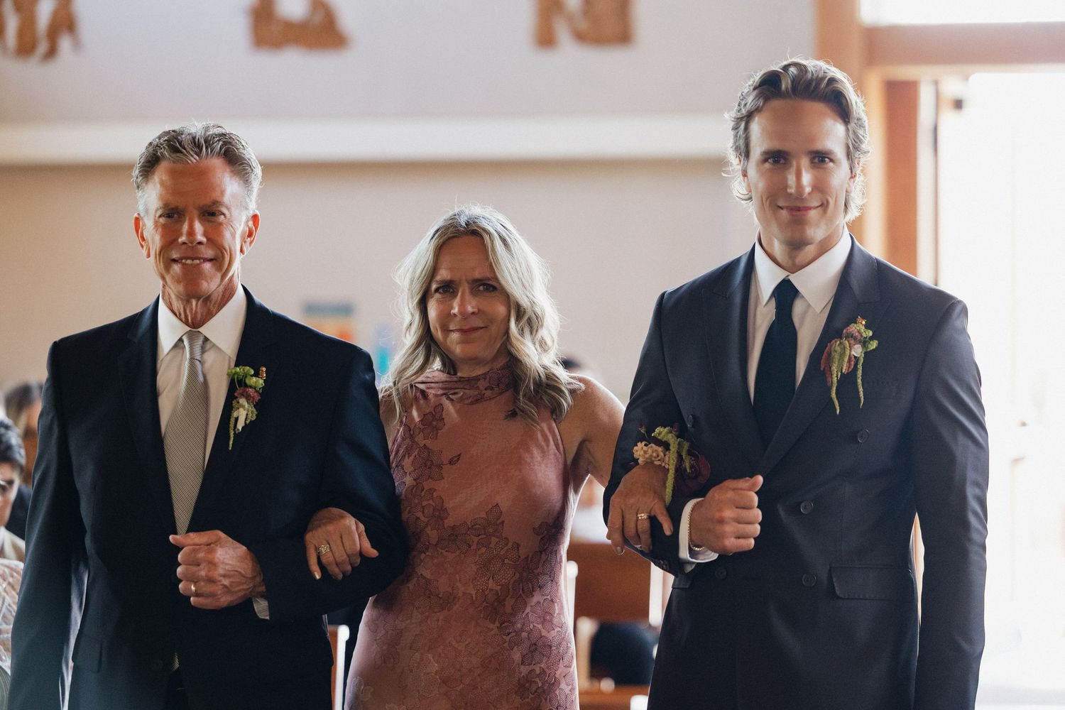 Two men in suits escort a woman in a pink lace dress down the aisle at a wedding ceremony.