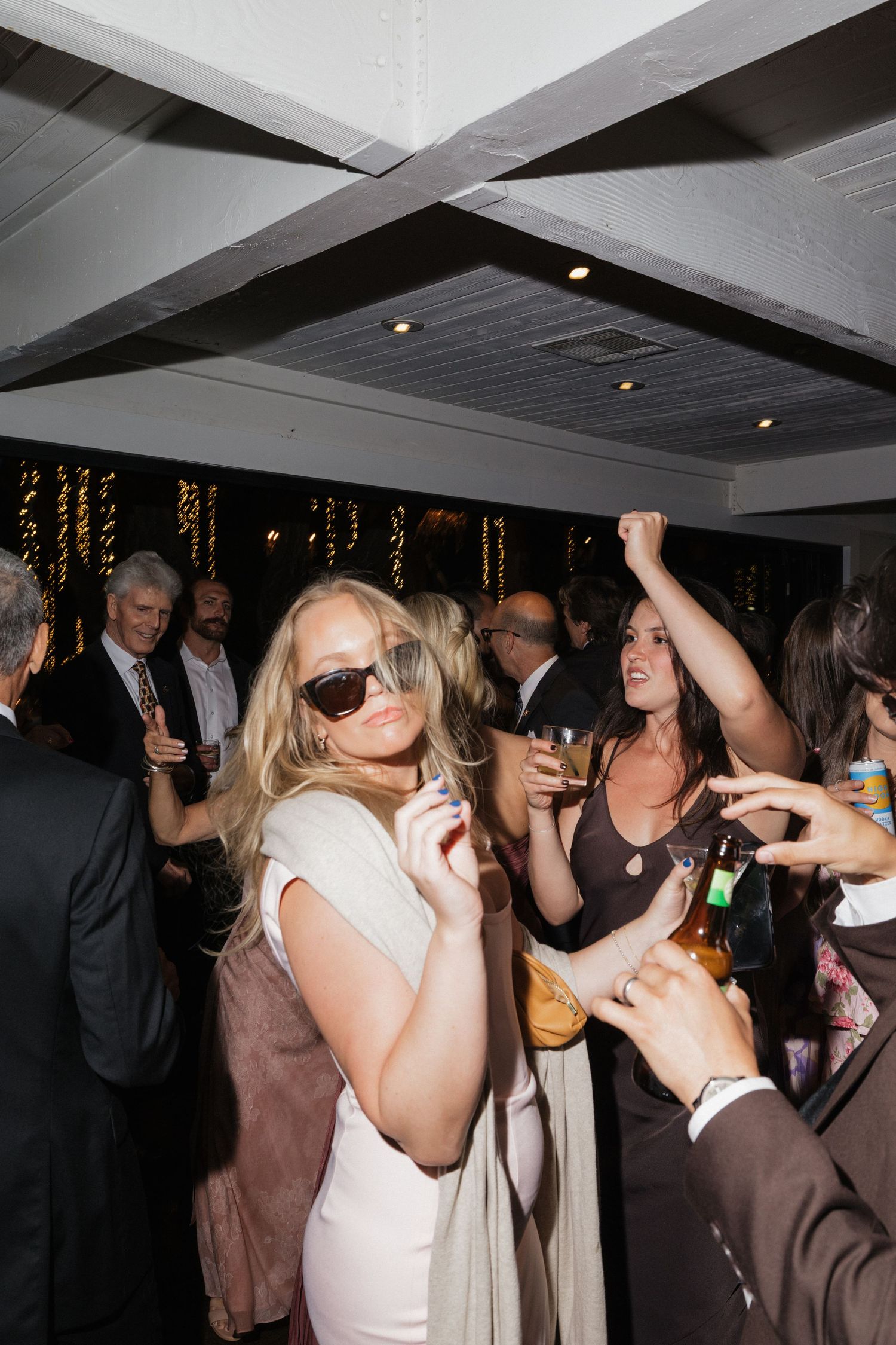 Wedding reception guests dance and celebrate indoors with sunglasses and drinks on the dance floor.