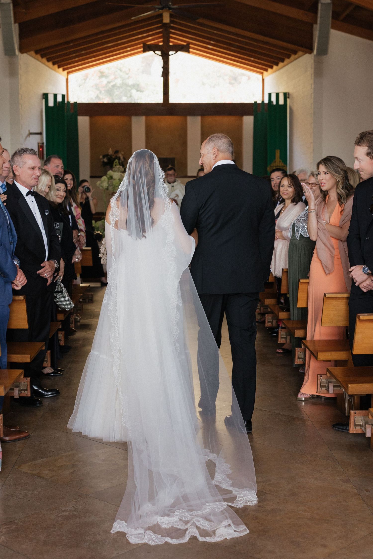 Bride walking down church aisle with escort, guests standing in pews, wearing white gown and long lace veil.