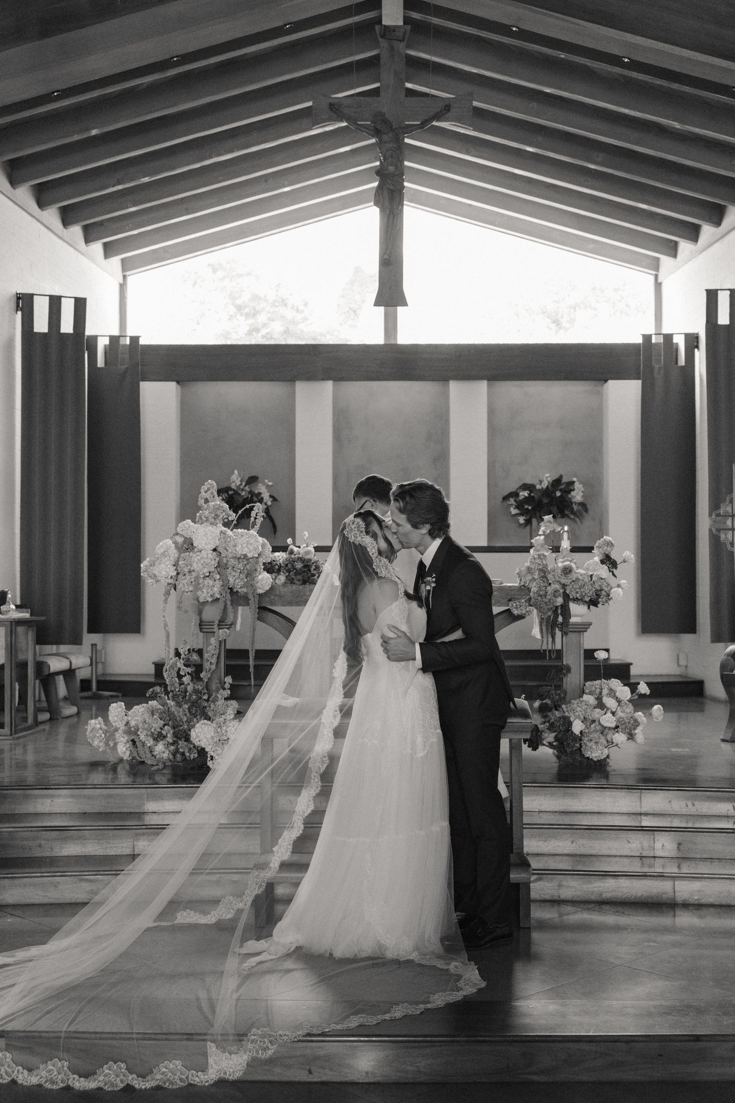 Bride and groom share first kiss at chapel altar with floral arrangements in black and white wedding photo.