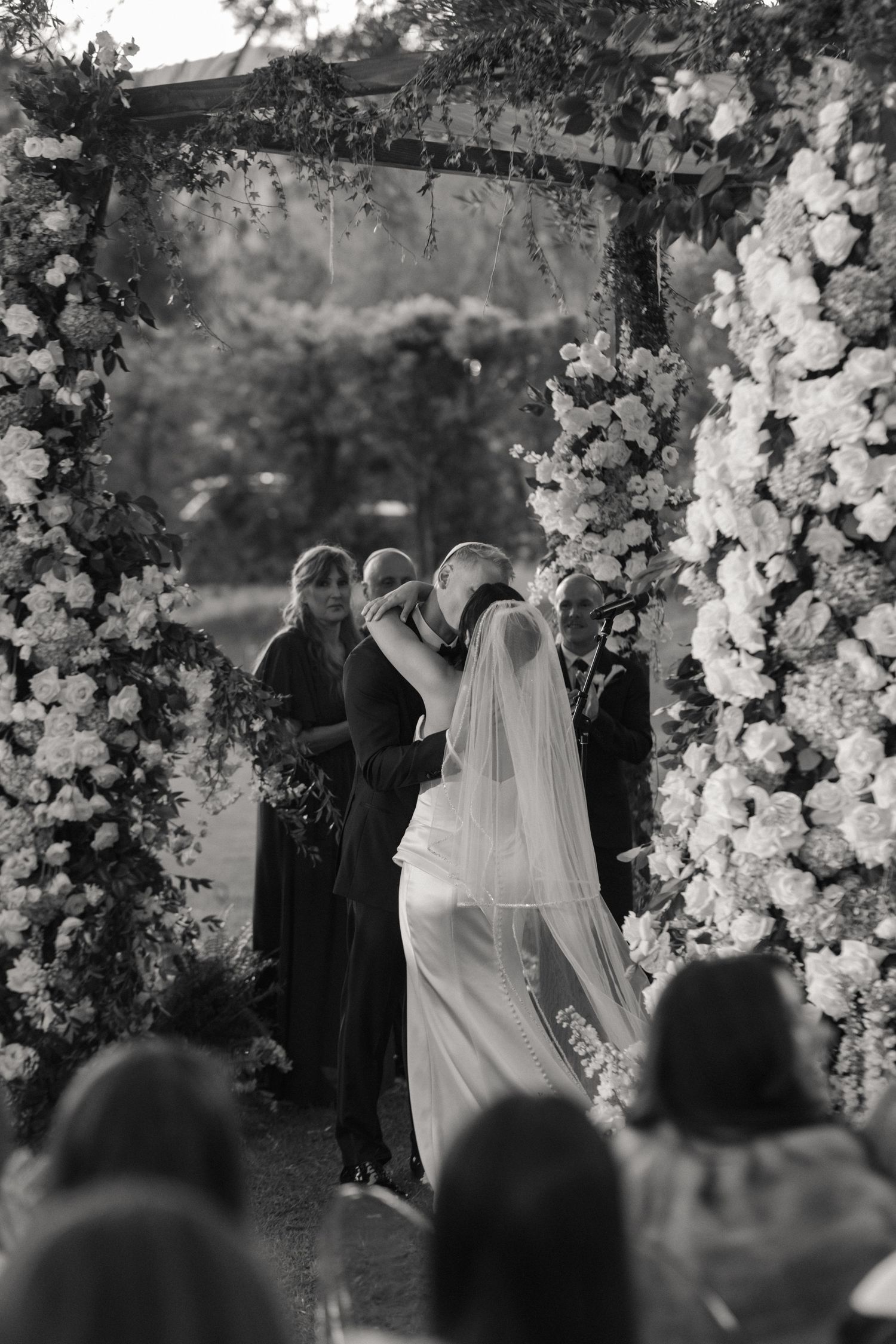 Black and white photo of bride and groom sharing first kiss under floral arch at outdoor wedding ceremony.