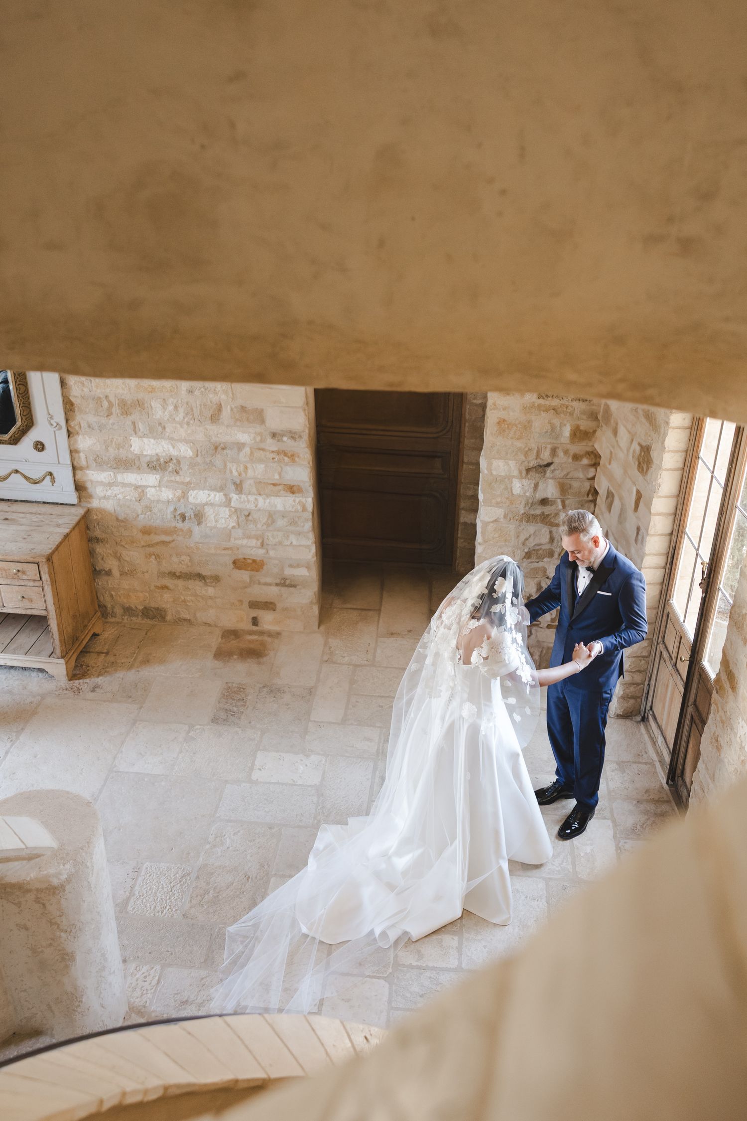 A couple in wedding attire dance together on an elegant stone staircase in a rustic building.