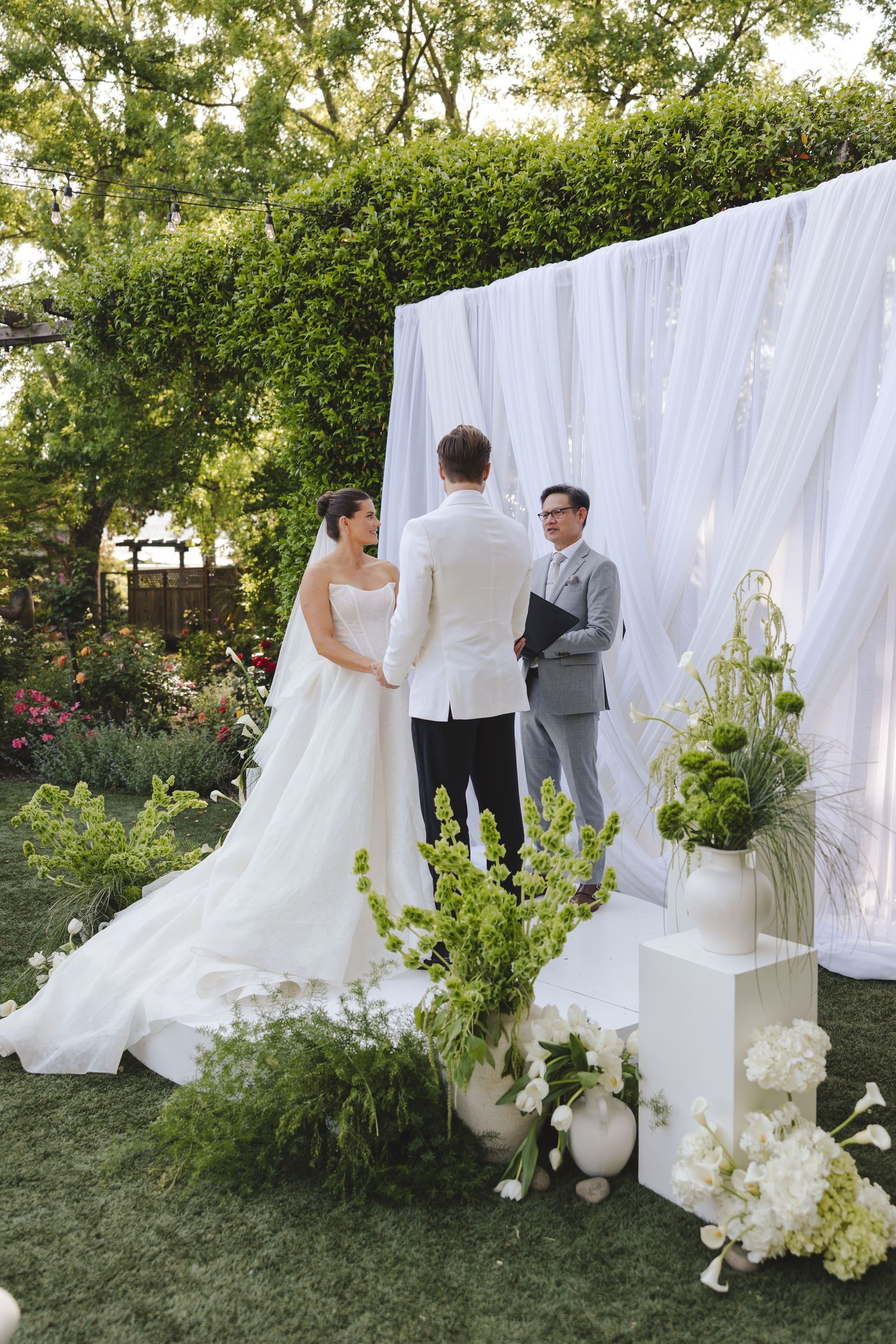 An outdoor garden wedding ceremony setup features elegant white drapery backdrop and floral arrangements on a grassy lawn.