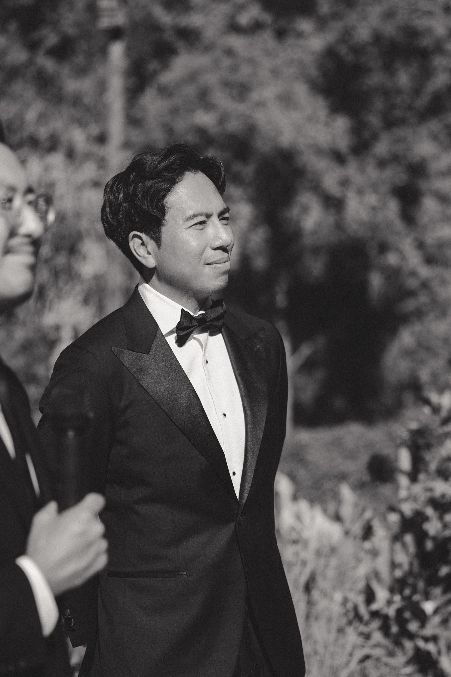 Groom in black tuxedo smiling and laughing during outdoor wedding photography session.