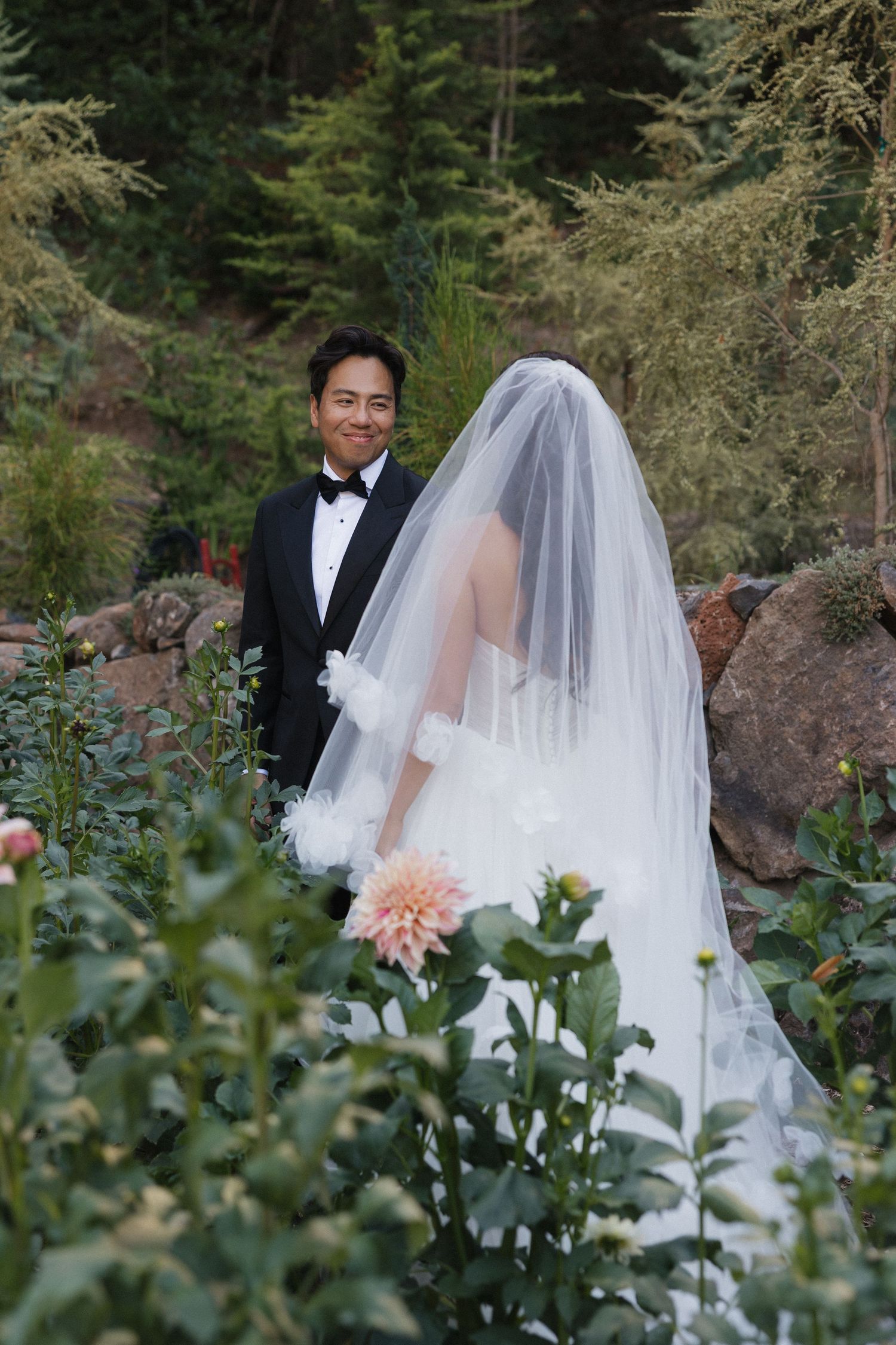 Wedding couple in formal attire walking through a garden with evergreen trees.
