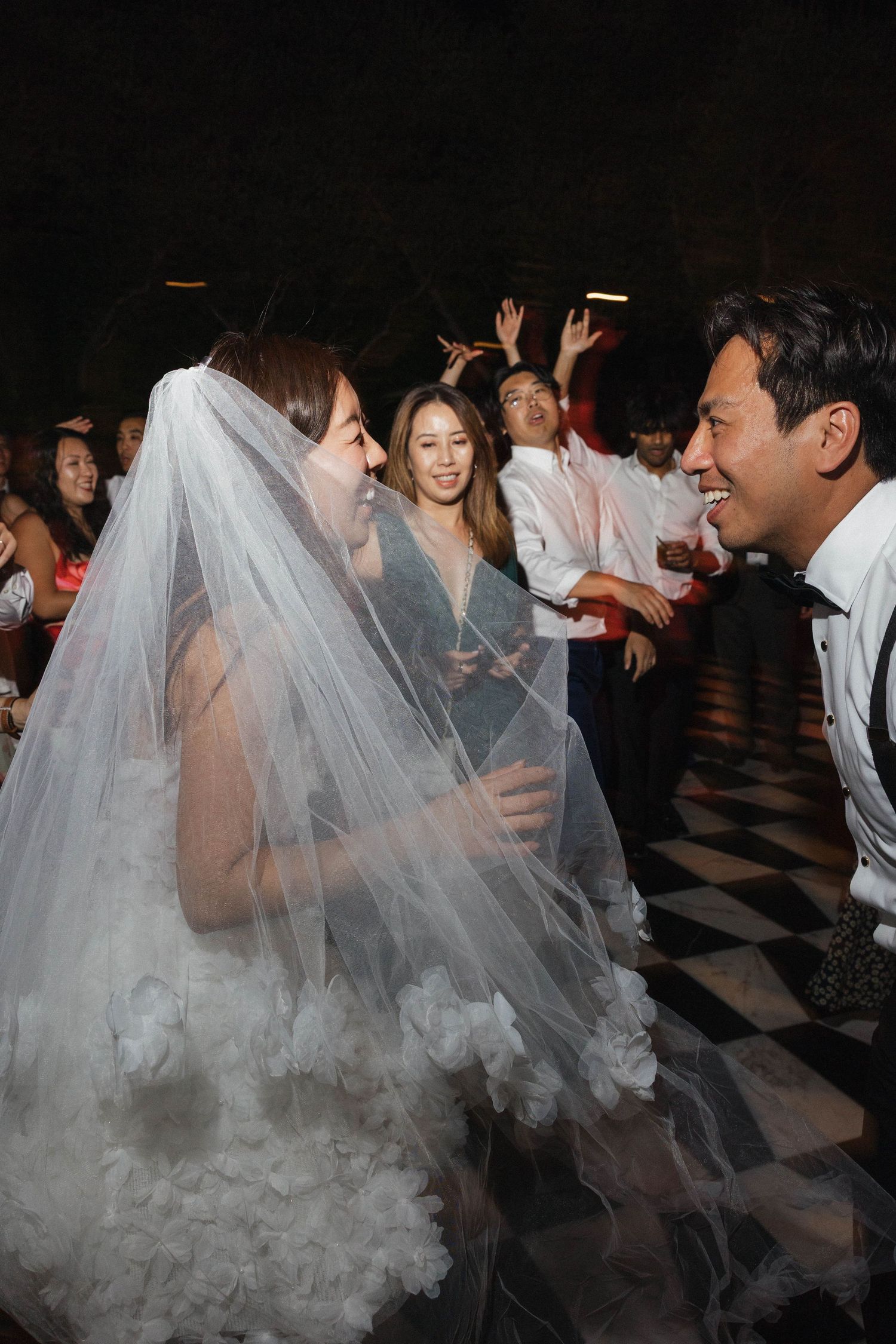 Joyful wedding celebration on a checkered dance floor with guests dancing and a bride in a flowing veil.