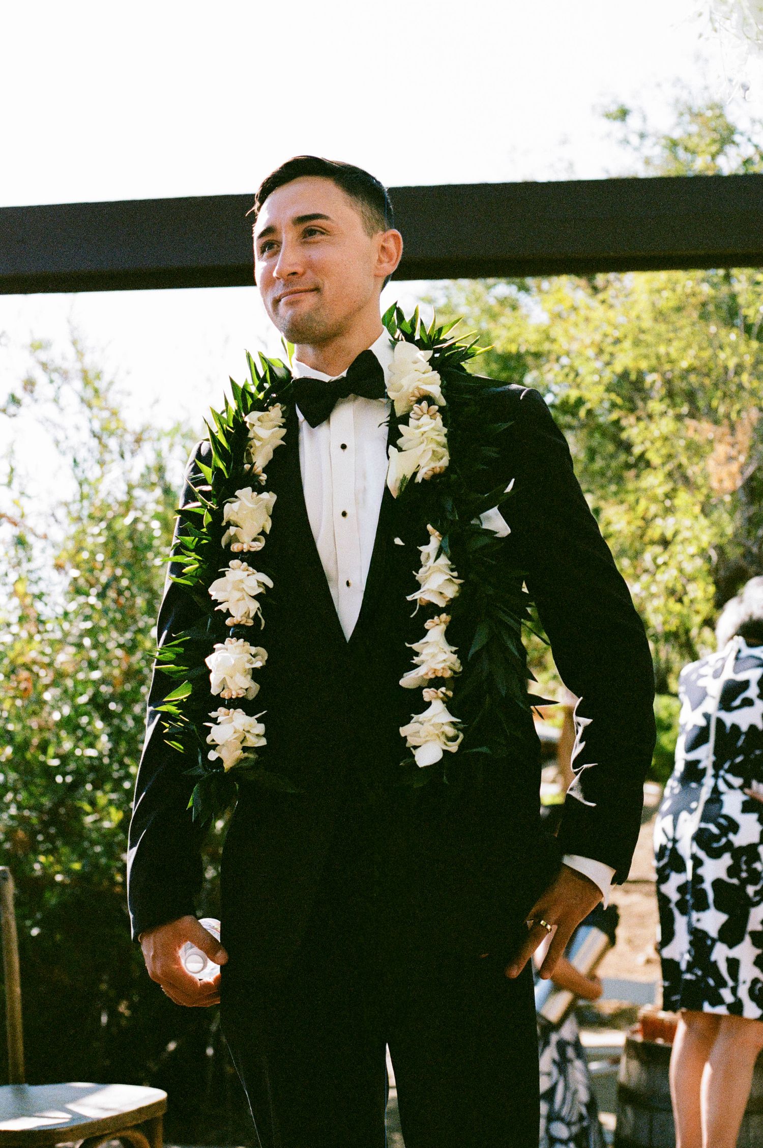 A wedding party with groomsmen in black tuxedos wearing white floral leis outdoors with natural lighting.