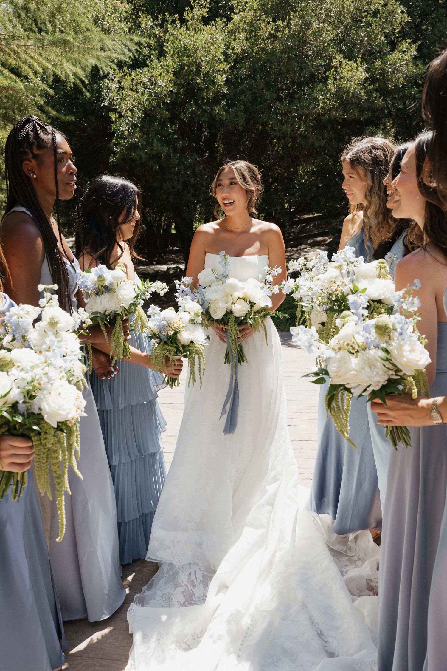 Bridesmaids in light blue dresses hold white bouquets while standing with bride in white gown outdoors.