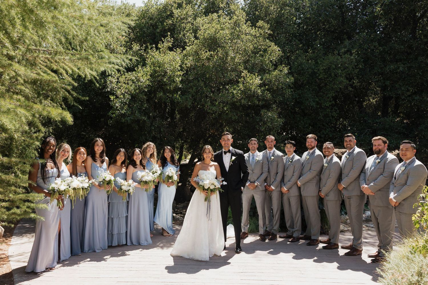 Large wedding party posed in a curved line wearing gray and white formal attire.