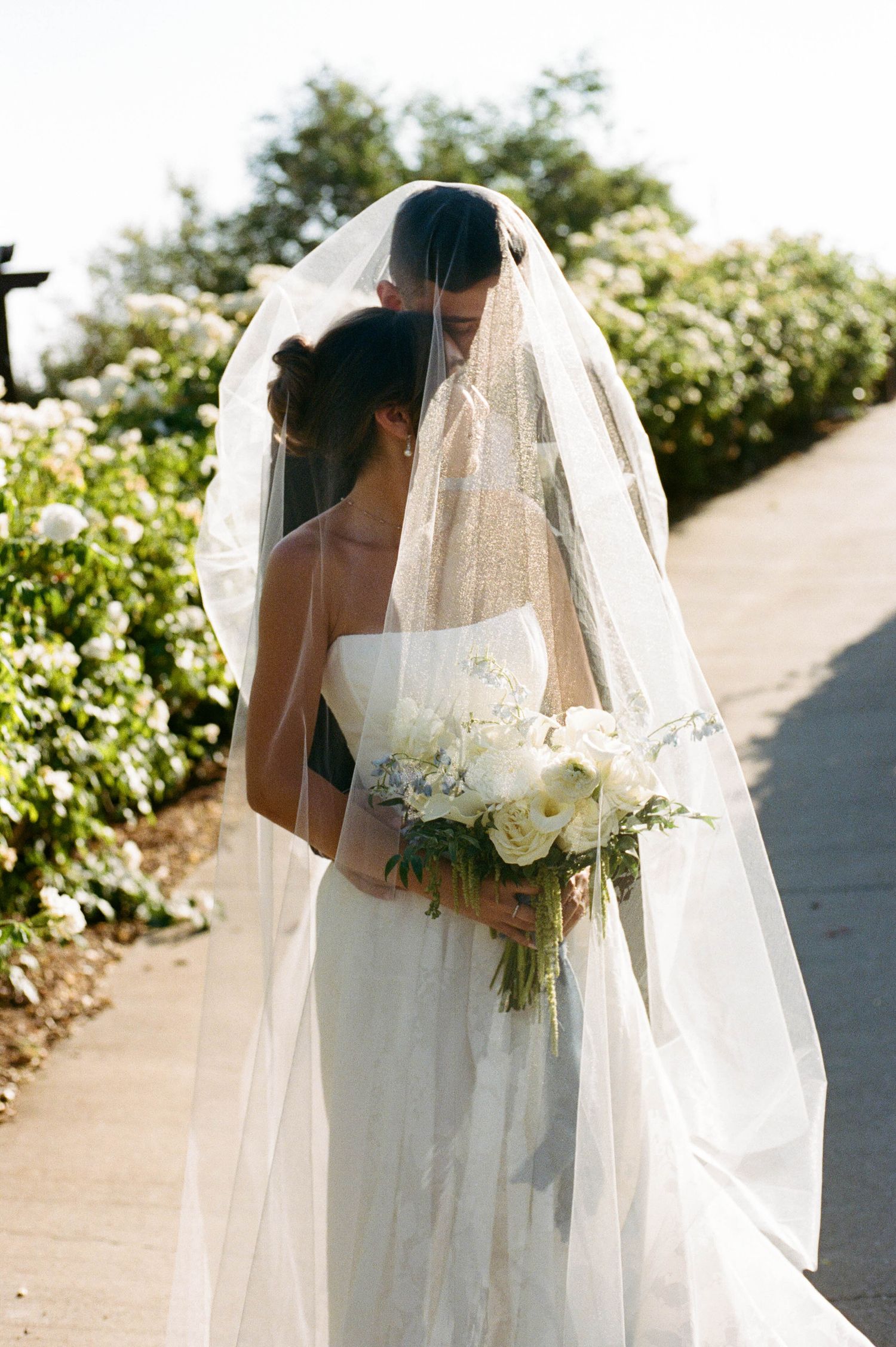 An elegant bridal veil and white bouquet against a sunlit garden backdrop.