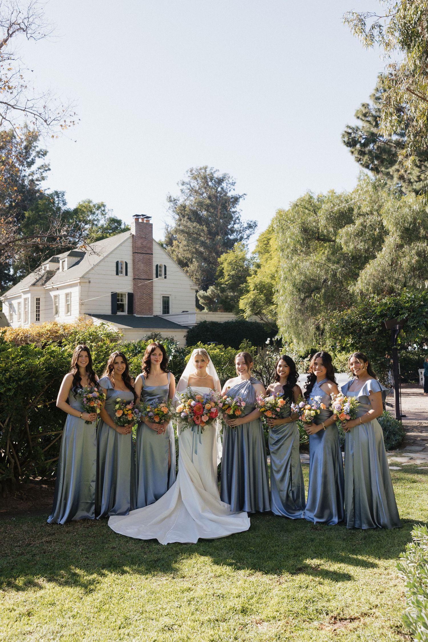 Wedding party in dusty blue bridesmaid dresses poses together on a sunny lawn with autumn-colored bouquets.