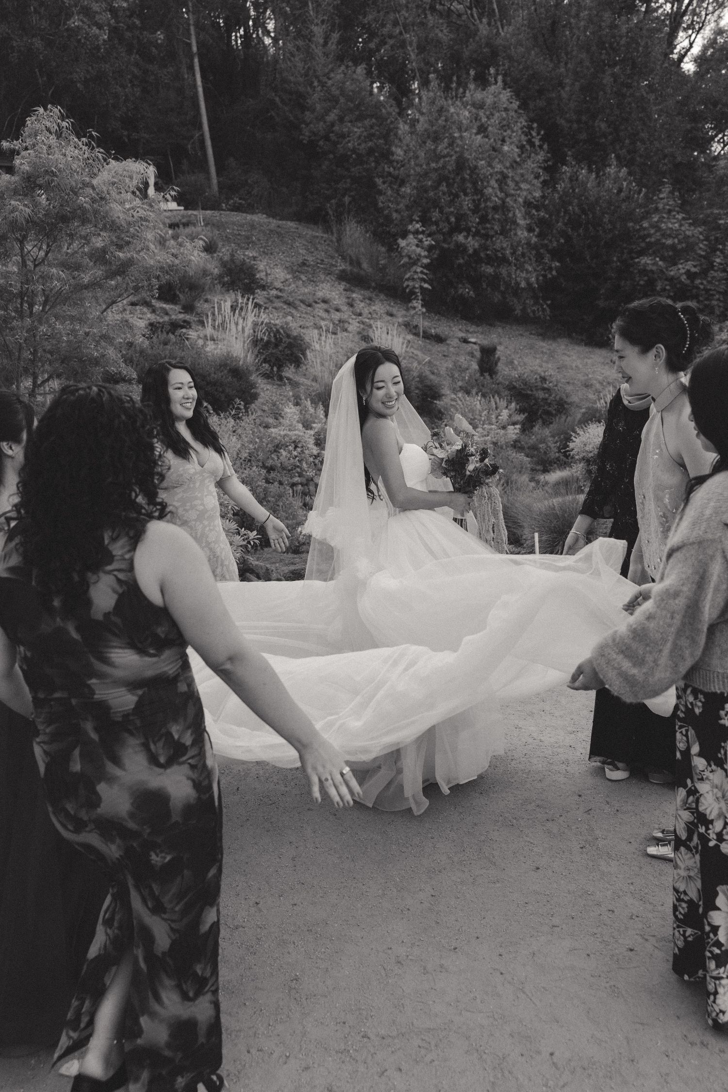 A bride in a white wedding dress gets help with her gown from wedding attendants on a path.