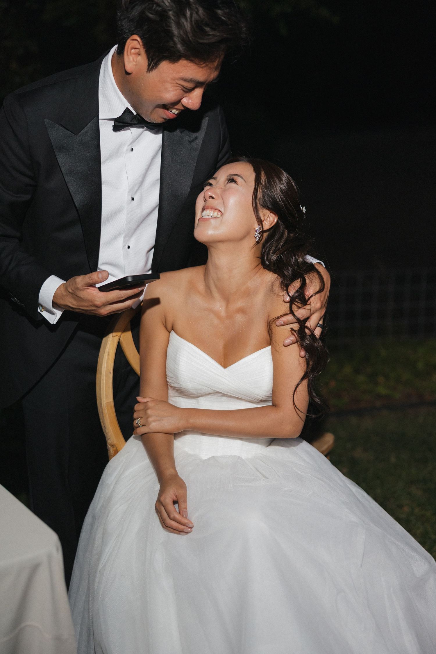 A bride in a white wedding dress laughs joyfully while looking up at her groom during an evening reception.